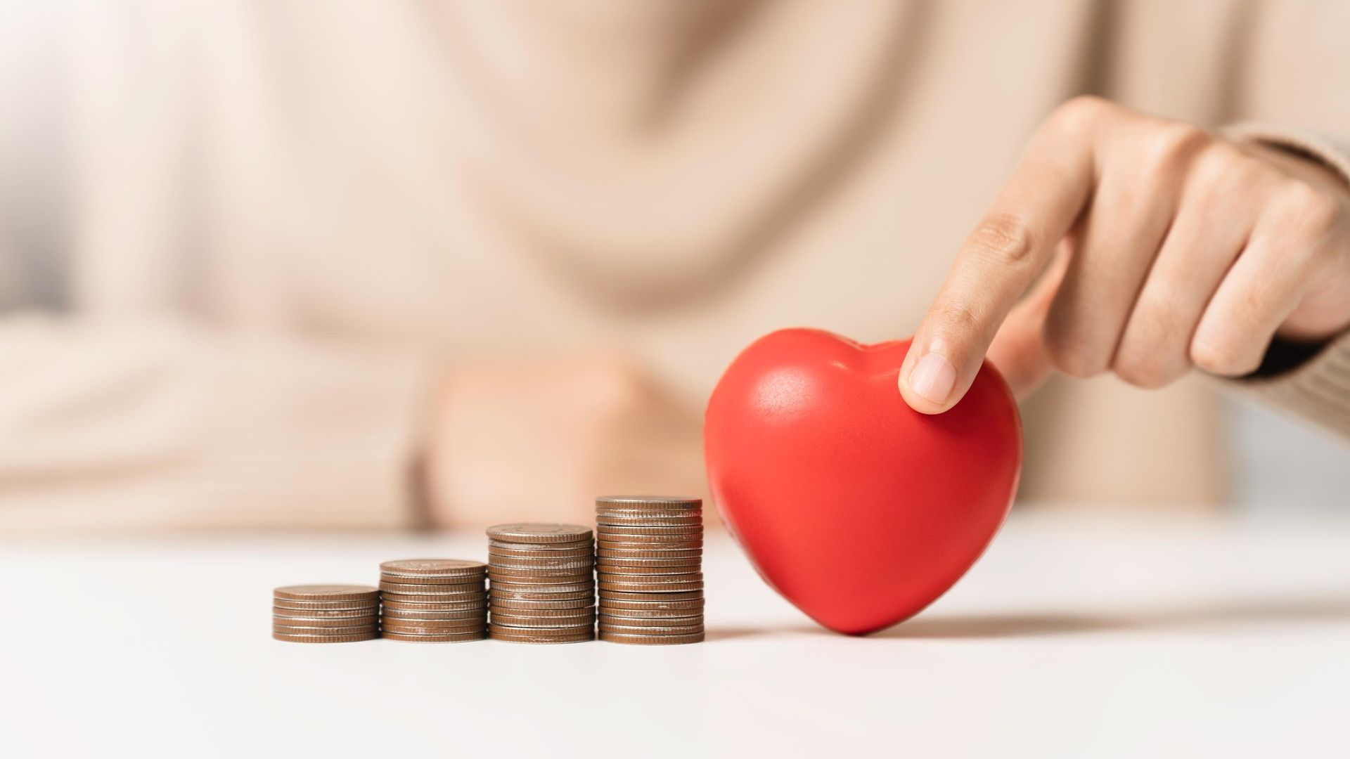 A person's finger touches a red heart next to stacks of coins, symbolizing charity.