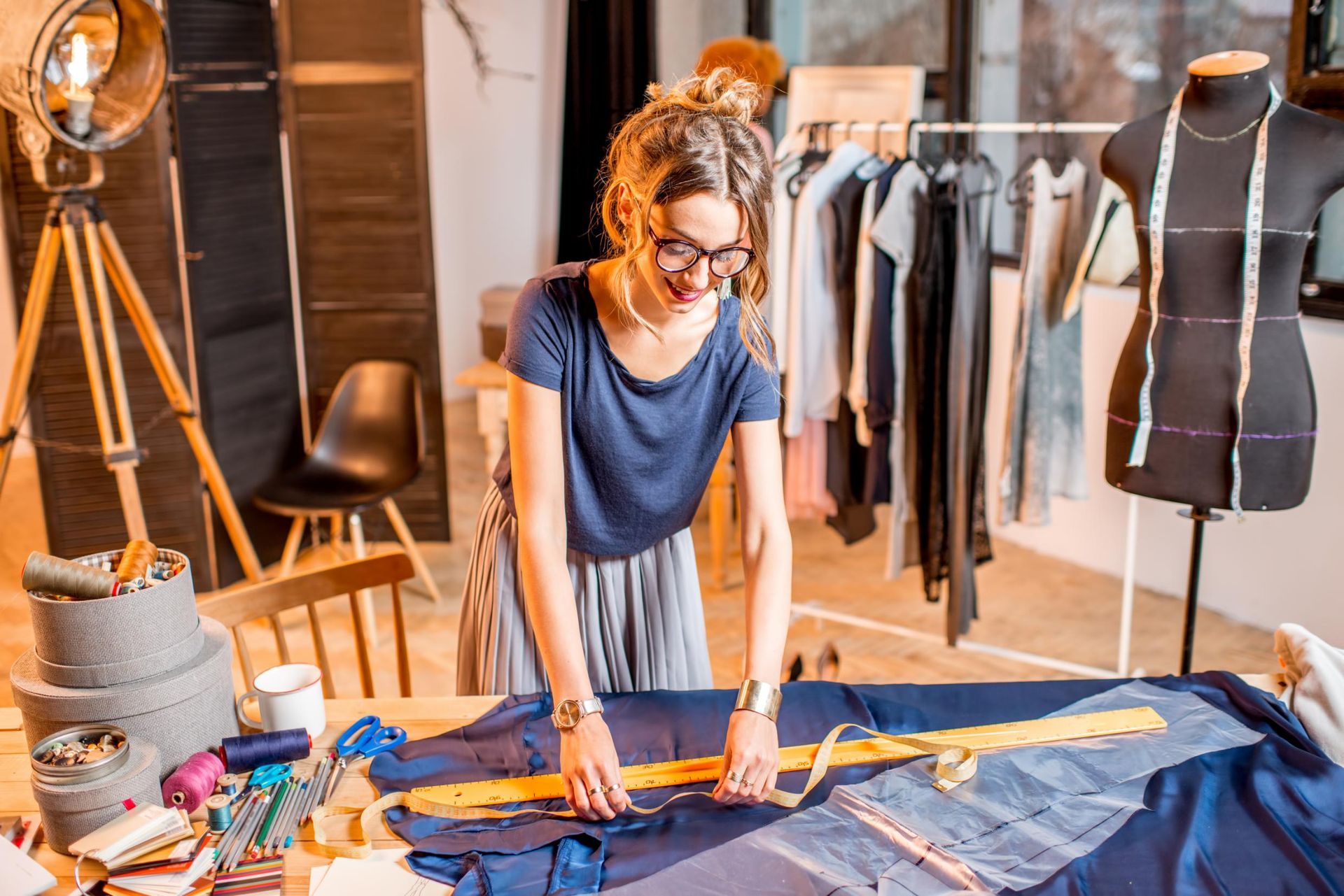 Woman in glasses measures fabric at a sewing table in a fashion studio, with clothes rack and mannequin.