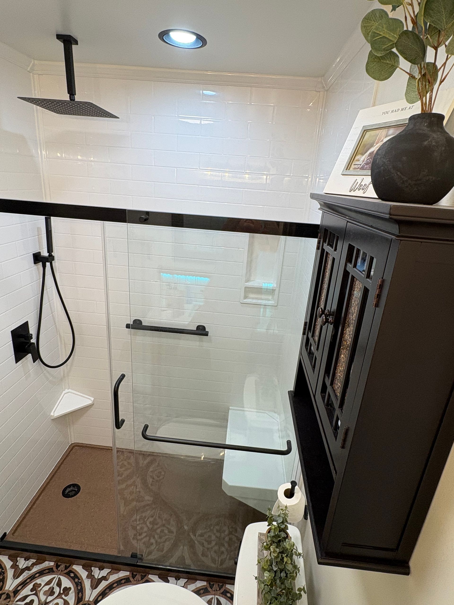 Bathroom with a black showerhead, glass door, and a brown patterned floor. A black cabinet sits on the wall.
