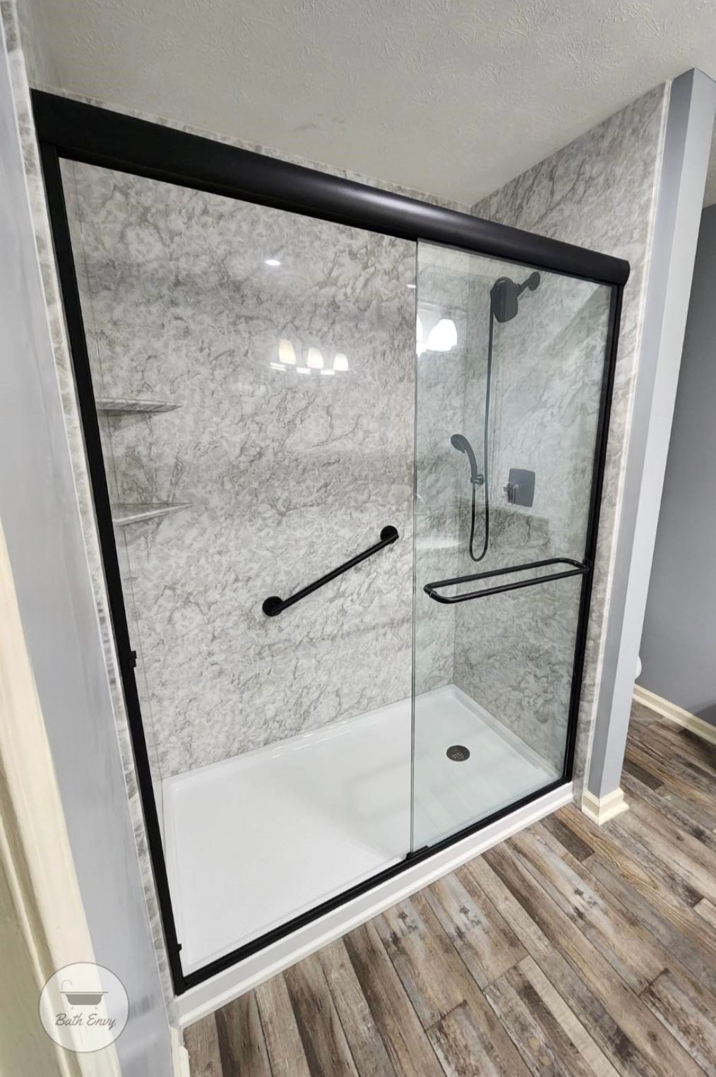 Black-framed shower with sliding glass doors, grab bars, and white base, set against gray textured walls.