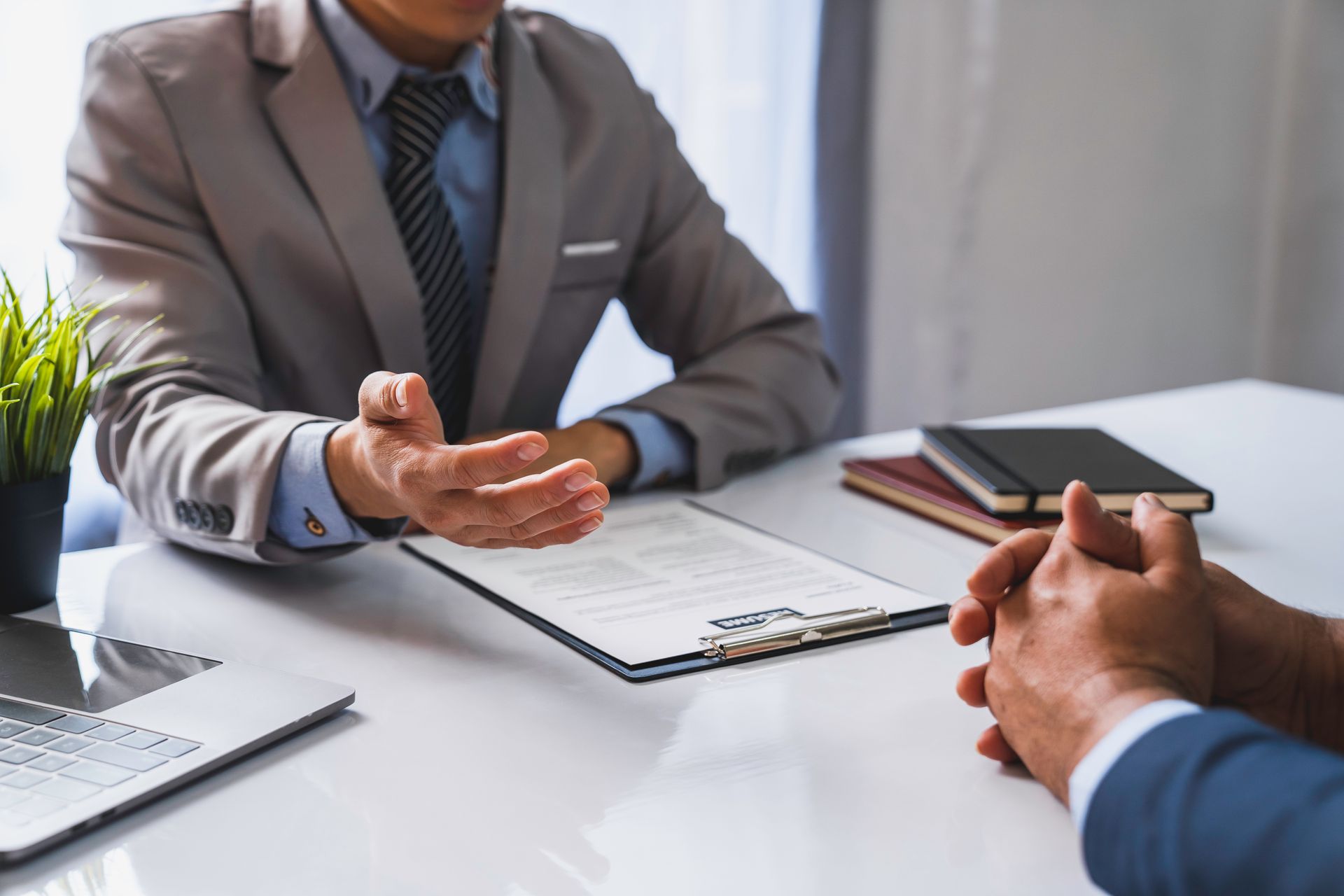 Two men in suits at a desk, one gesturing while speaking, the other listening attentively; office setting.