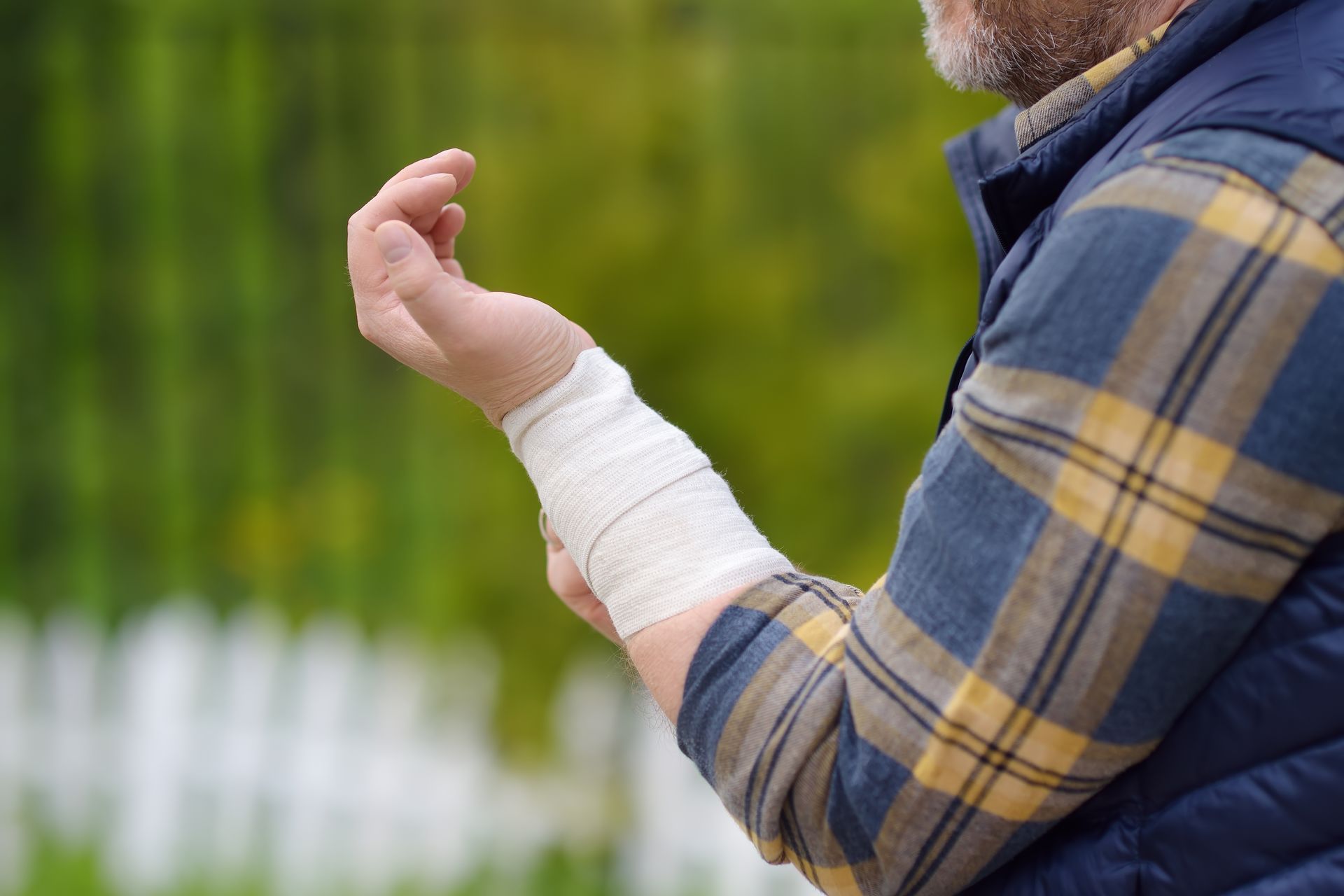 Man with wrapped wrist, wearing blue plaid shirt and vest, outdoors. 