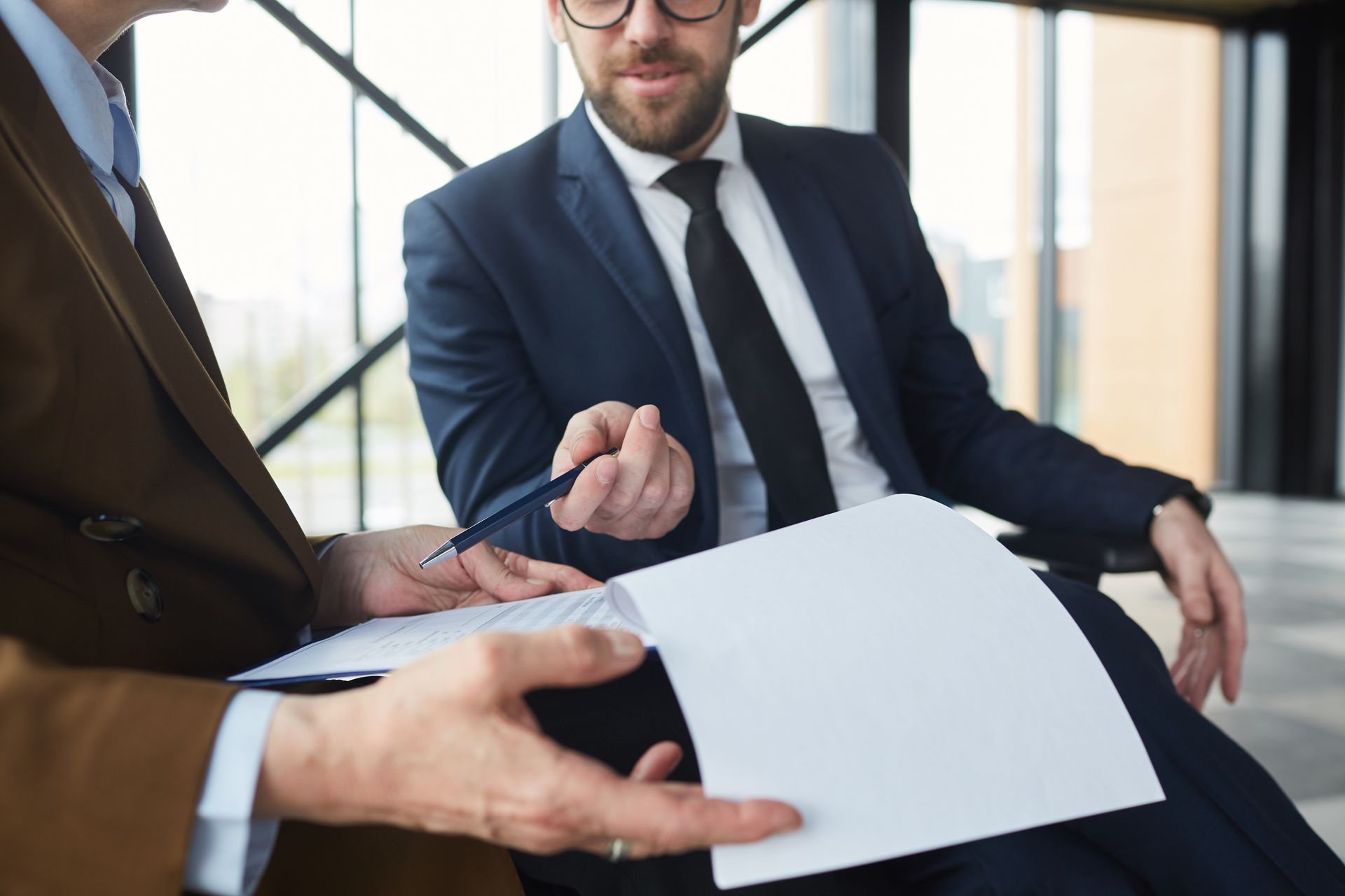 Two men in suits reviewing documents, one gesturing with a pen, in an office setting.