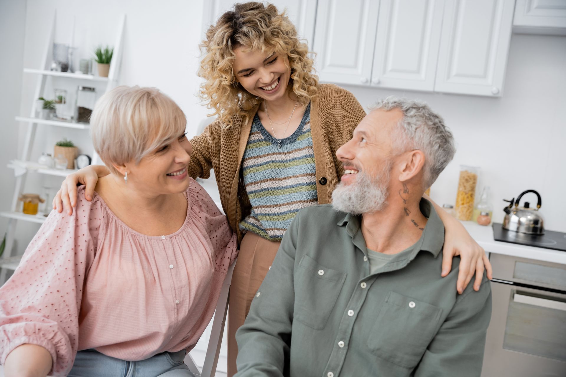 Woman with curly hair smiles at her parents in a kitchen, arm around her mother's shoulders.