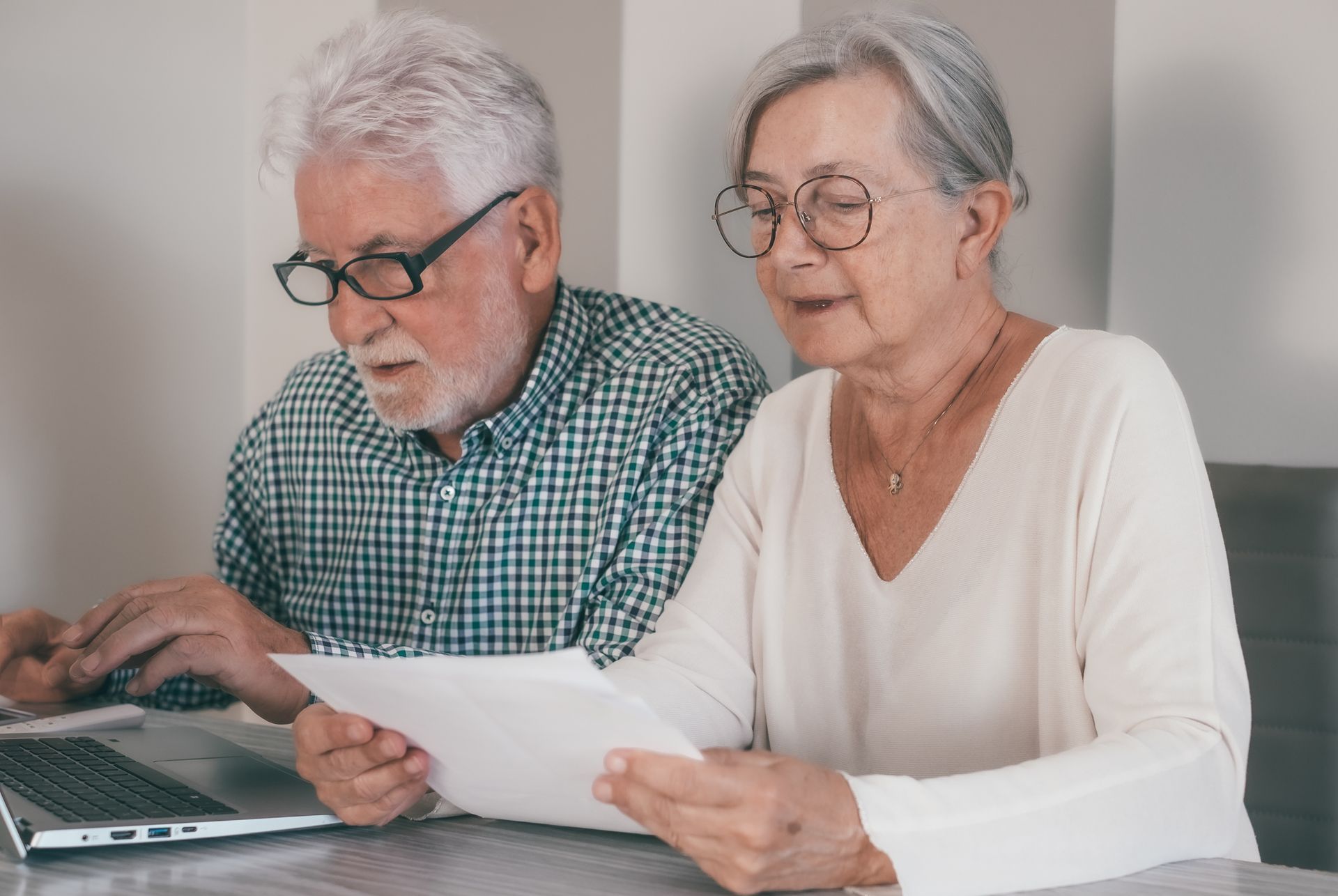 An elderly couple reviews documents near a laptop indoors, both wearing glasses and focused, with neutral expressions.