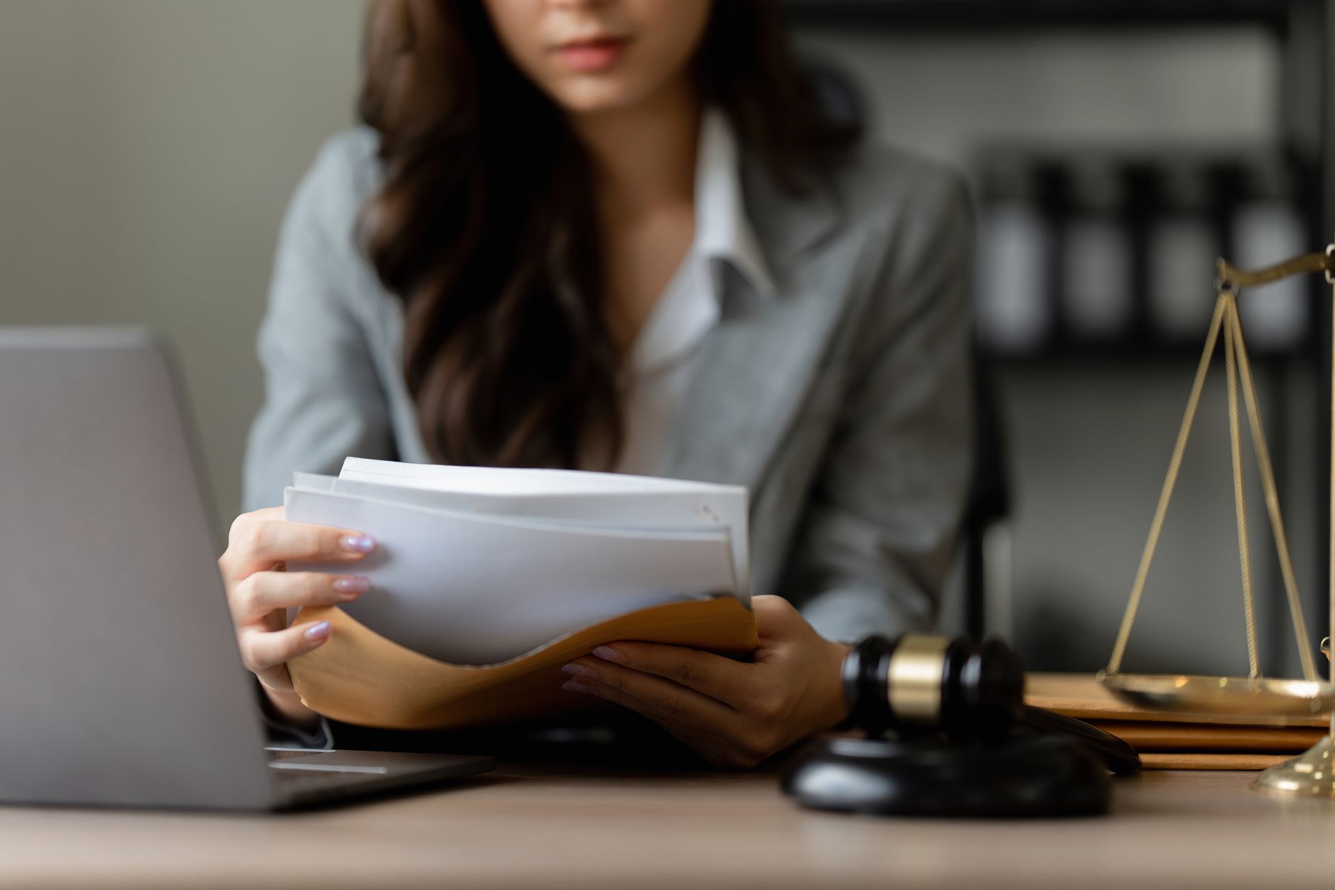 Woman in suit examining documents, with gavel, scales, and laptop on desk.