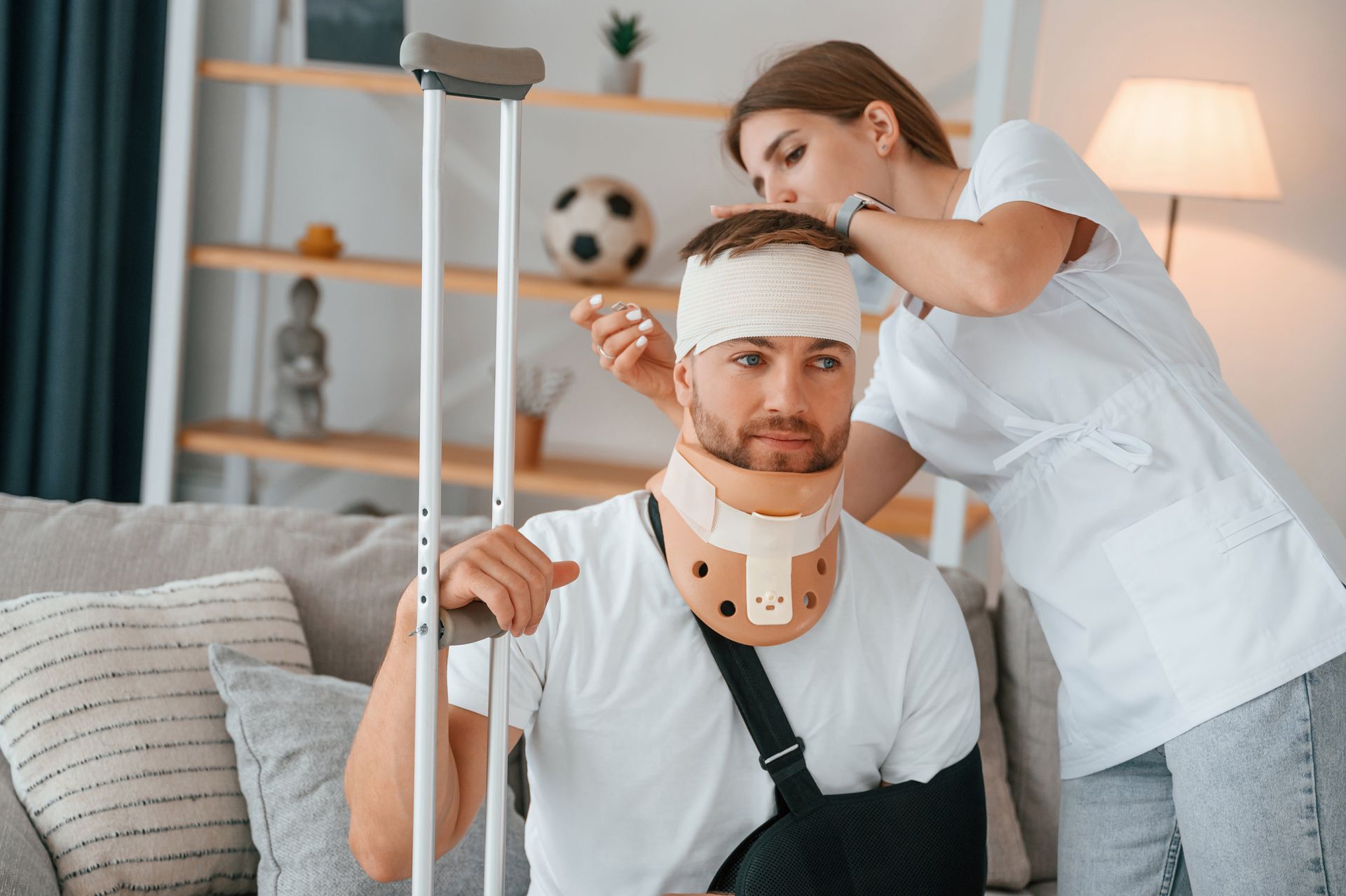 Woman adjusting bandage on man's head, wearing neck brace and arm sling, using crutches, sitting indoors.