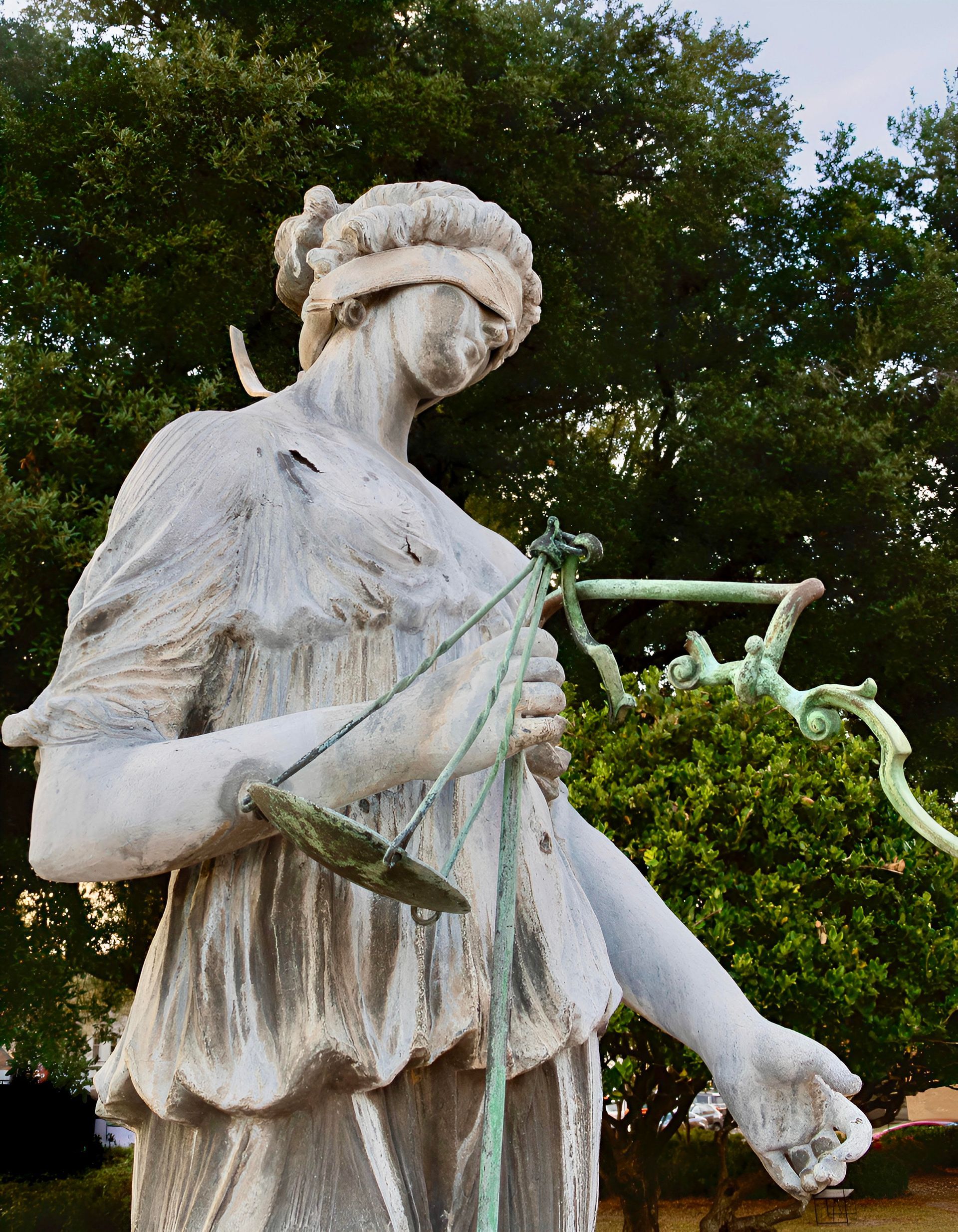 Statue of Lady Justice, blindfolded, holding scales. Outdoors, with green foliage background.