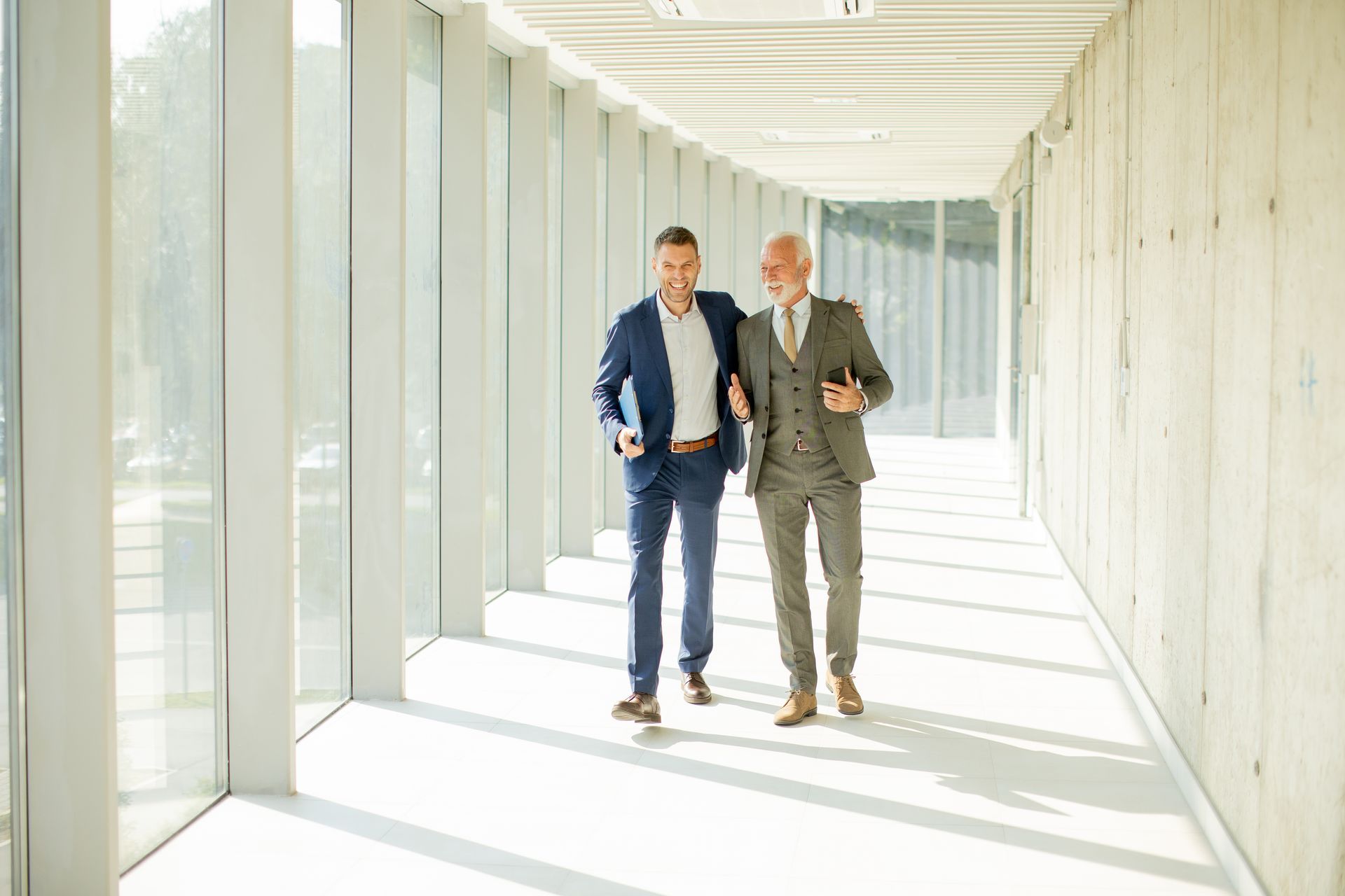 Two men in suits walking and talking in a bright hallway with windows.
