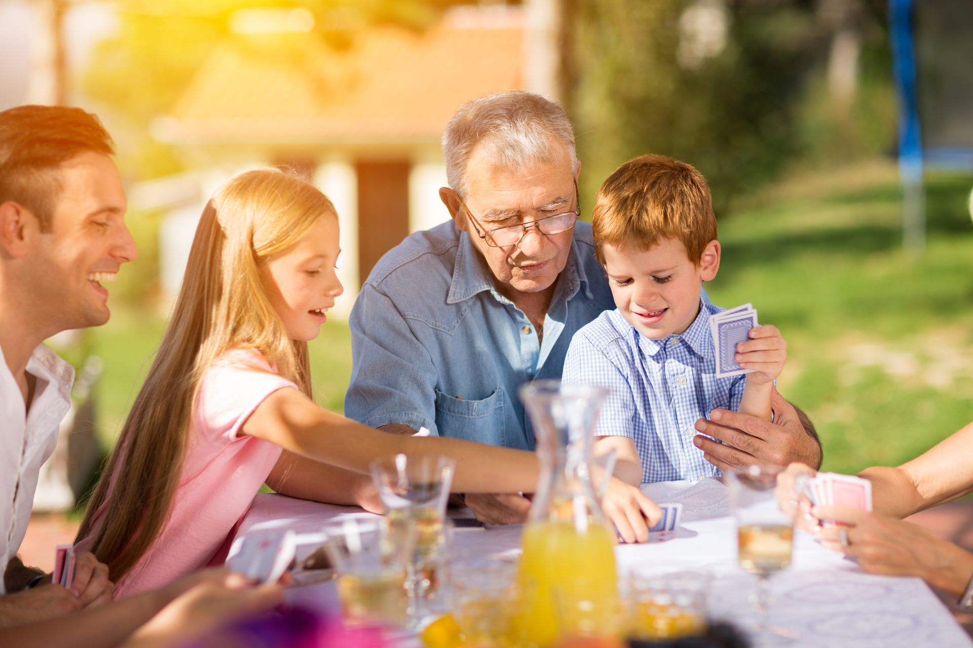 Family playing cards outdoors at a table. Child, adult, and older person interact. Sunny day.