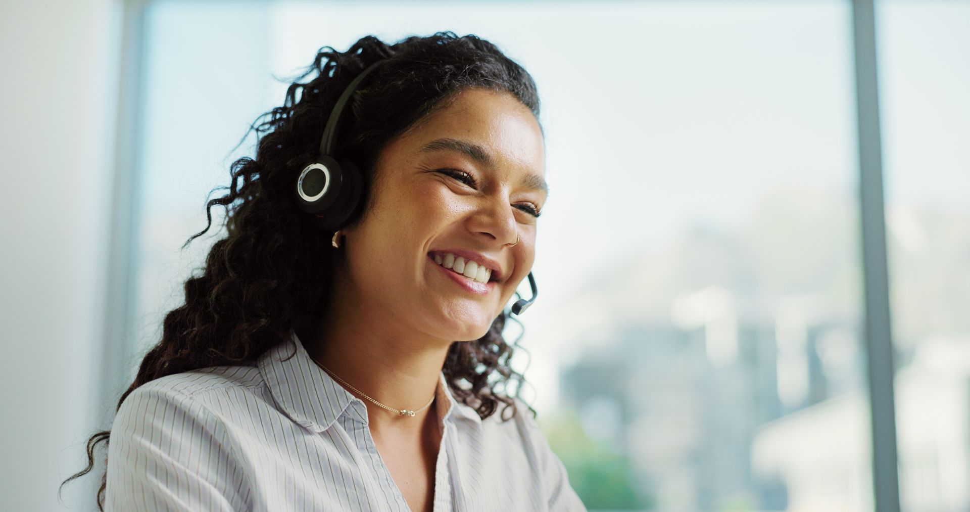 Woman wearing headset smiles brightly in a well-lit office setting.