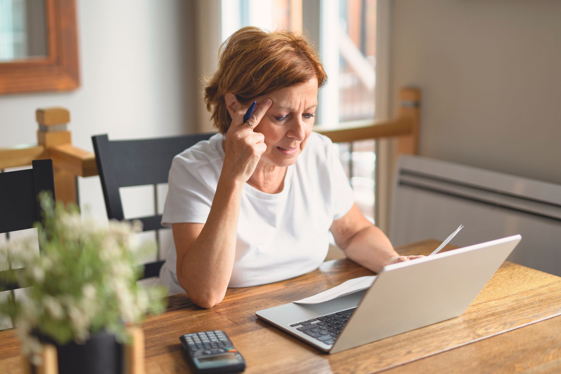 Woman at table, using laptop, looking stressed, hand on forehead, calculator nearby.