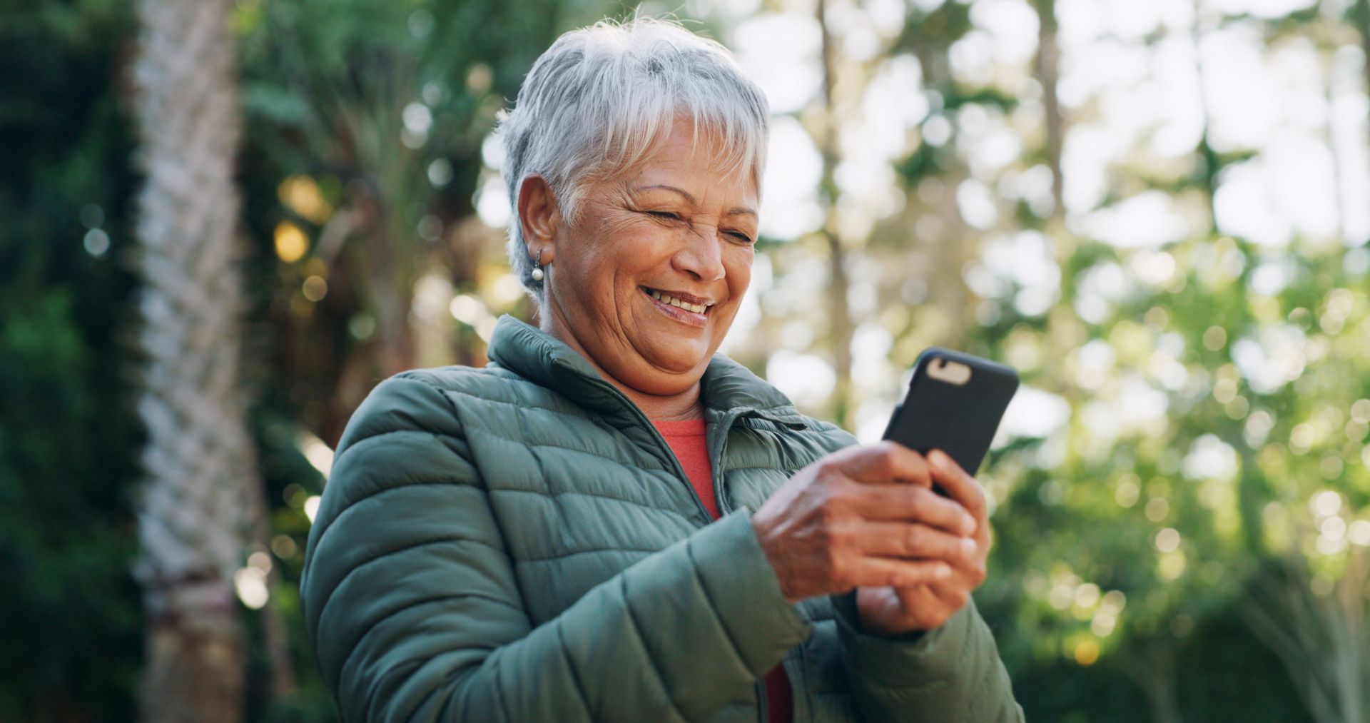 Woman smiles while looking at a phone outdoors, wearing a green jacket.