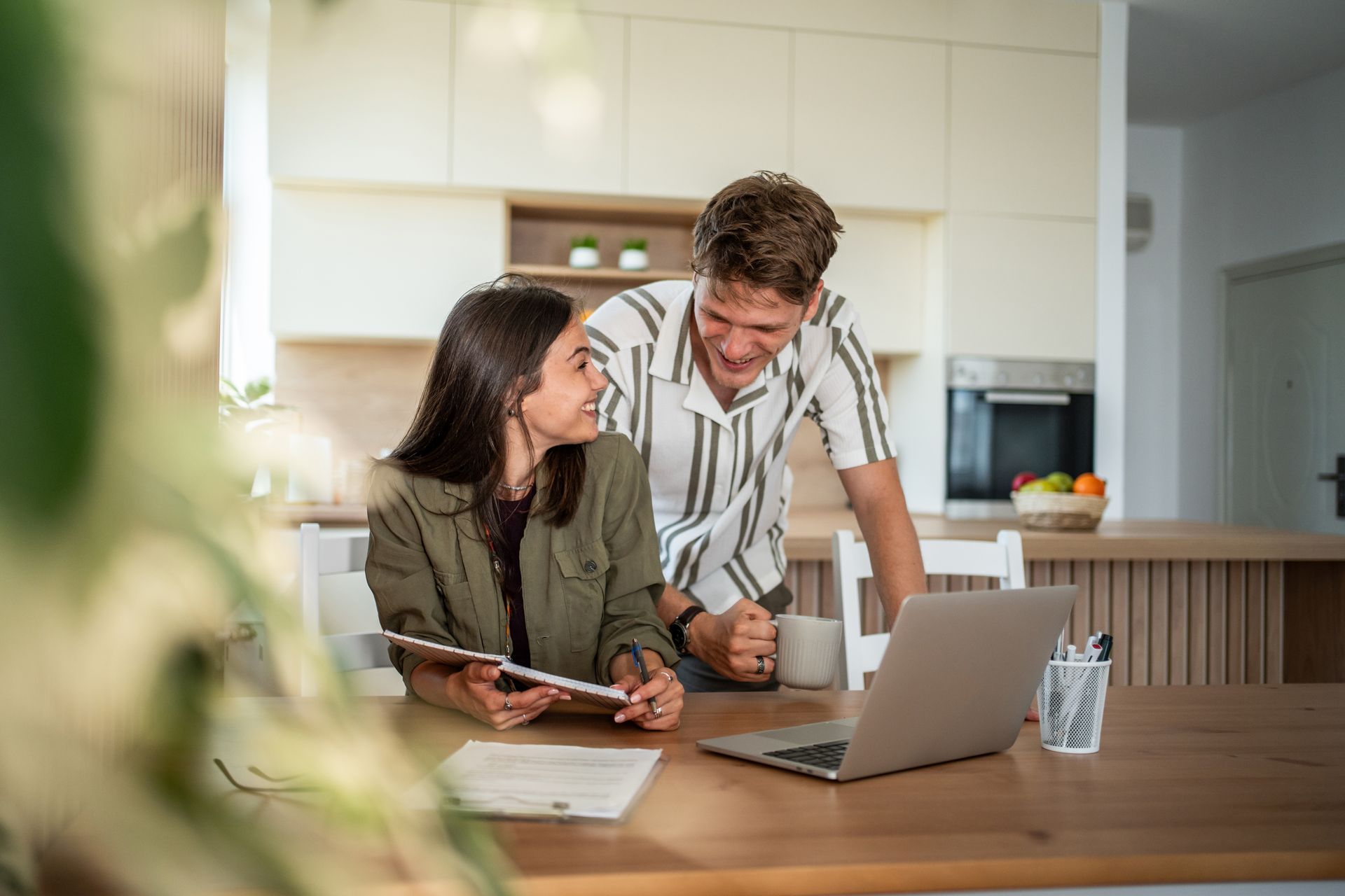 Couple looking at laptop and tablet at kitchen table, smiling, discussing finances.