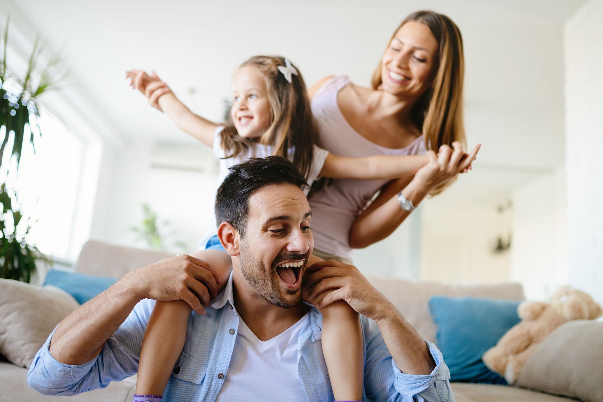 Family in a living room, smiling and playing. The father has the child on his shoulders, mother holds their hands.