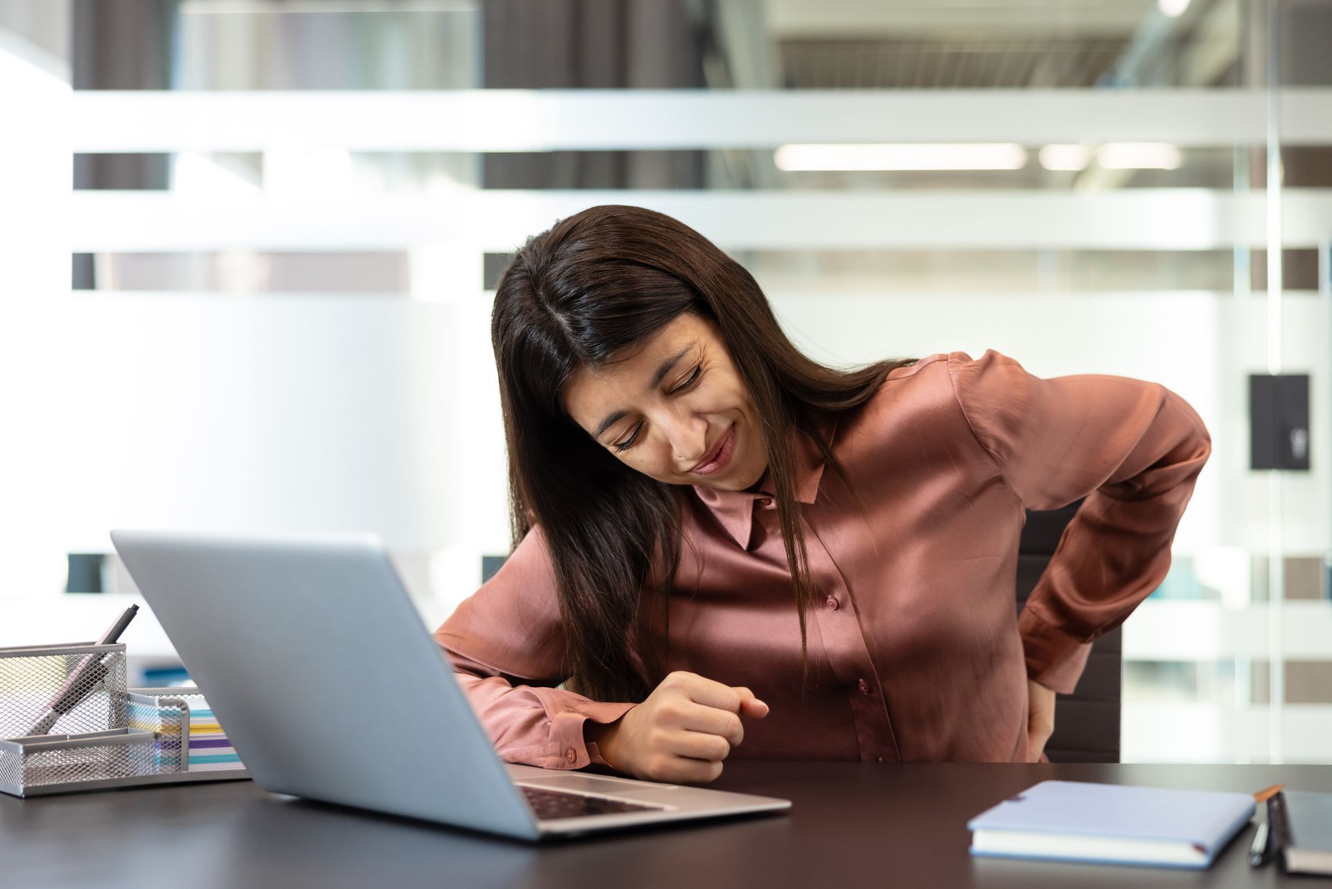 Woman at desk, holding her lower back with a grimace, next to a laptop and notebook in an office setting.