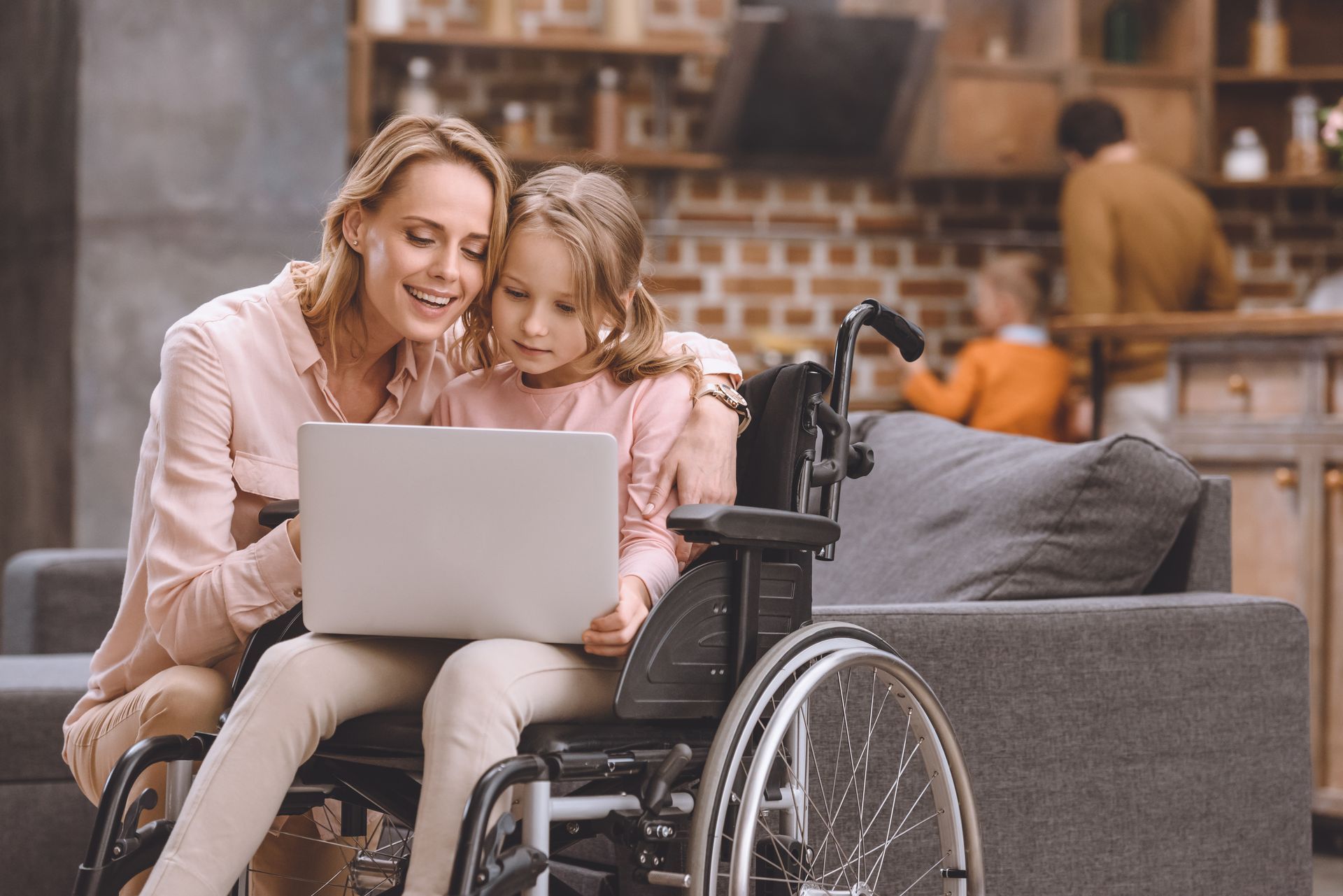 Woman and girl in wheelchair looking at laptop on a sofa; family in background.