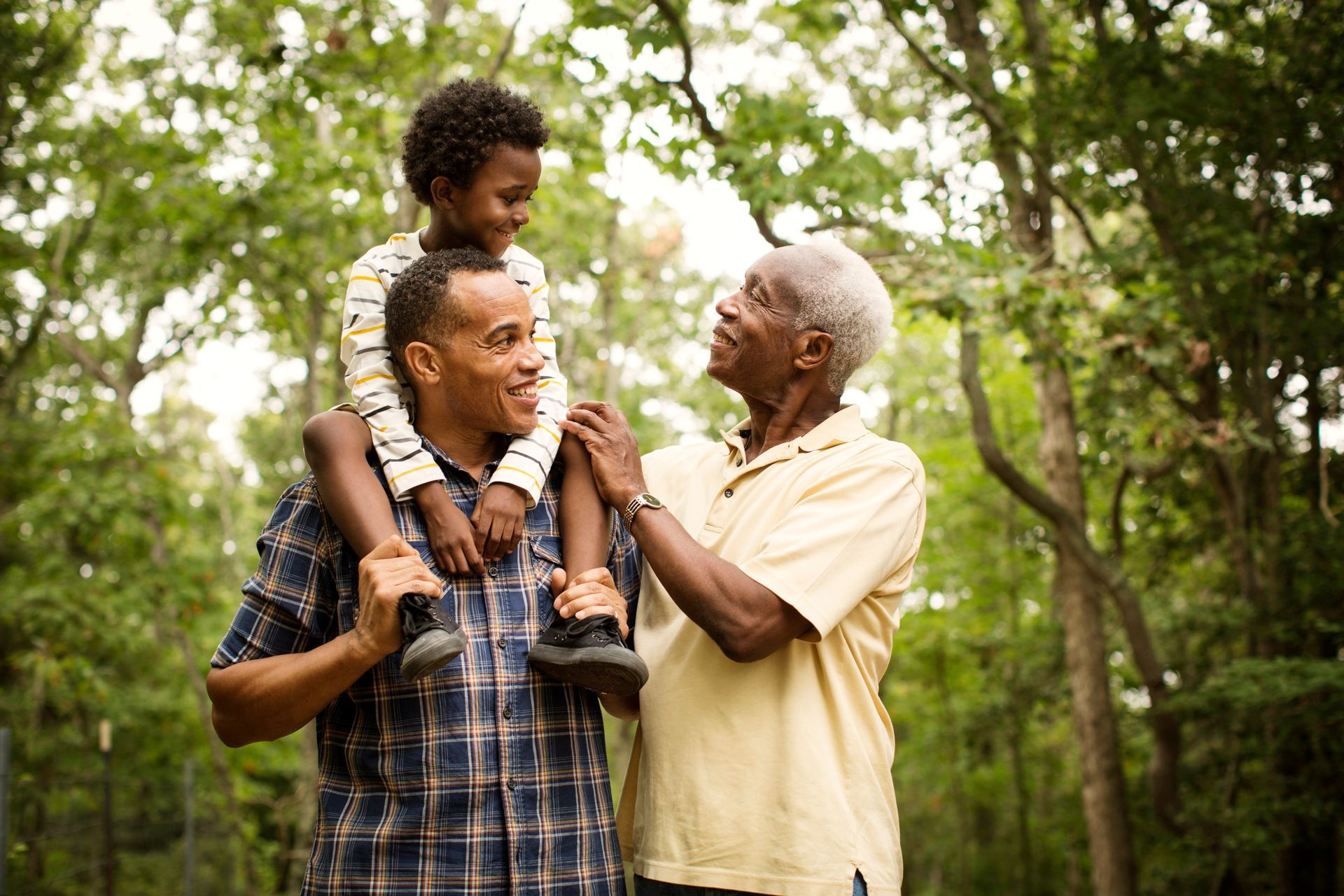 Man carries child on shoulders, smiles at older man, outdoors among trees.