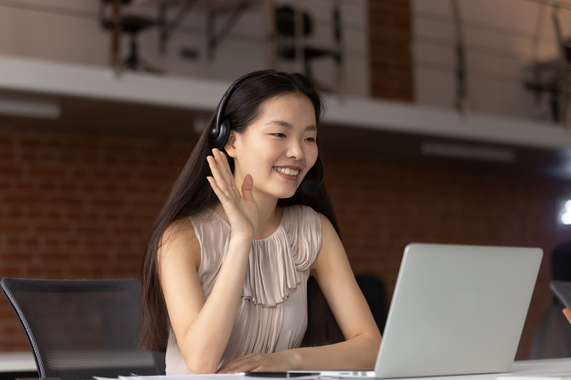 Woman with headset waves while on a laptop video call in an office.