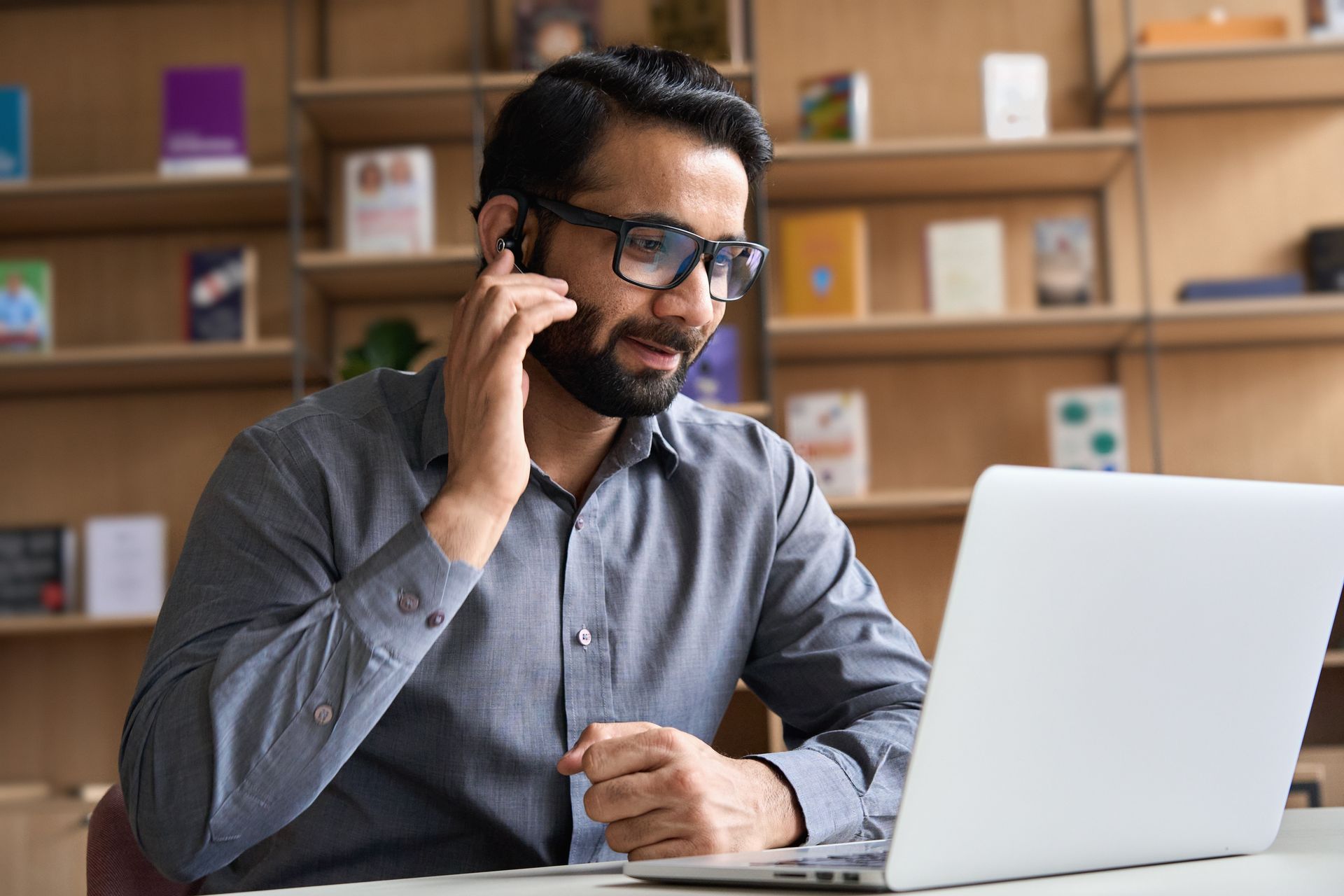 Man in glasses, using laptop, speaking on headset, in front of a bookshelf.