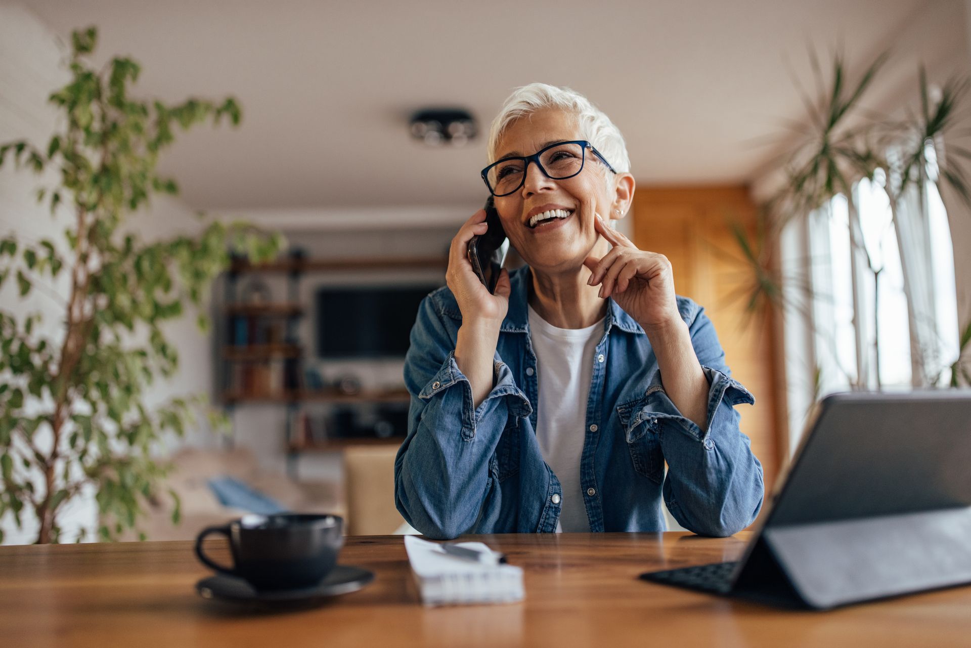 Woman with glasses smiles while talking on a phone, sitting at a table with a laptop, coffee, and plants.
