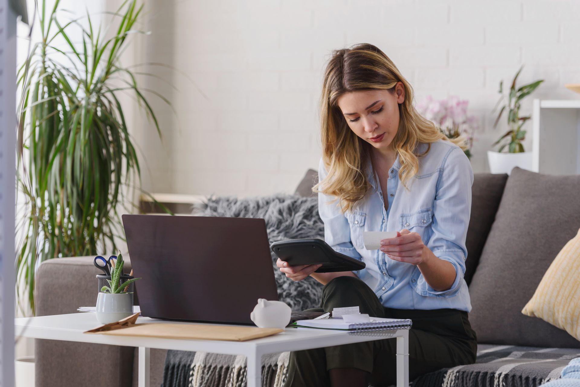 Woman calculating finances on a laptop and calculator, sitting on a couch in a living room.