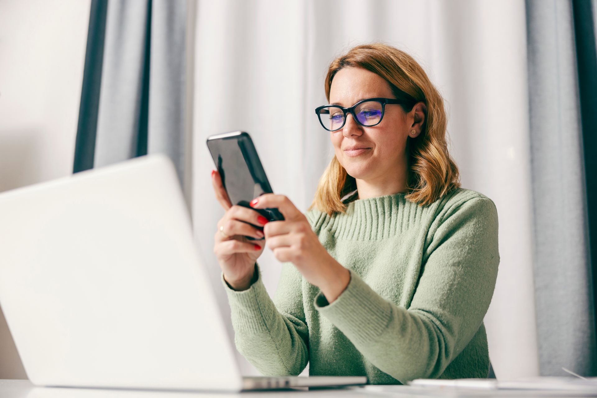 Woman in glasses holding phone, looking at screen, working at laptop.