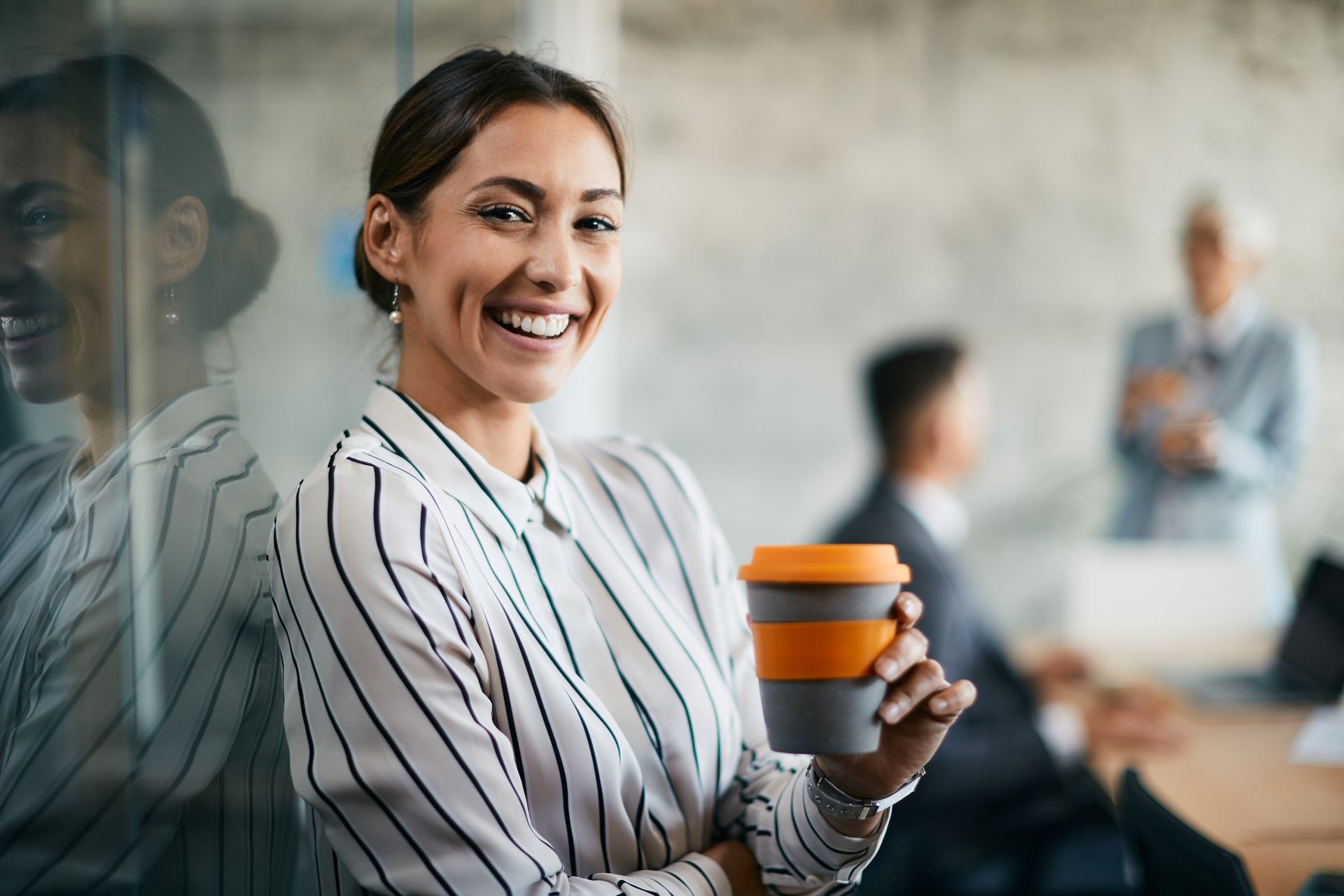 Woman with coffee cup smiling in office, leaning against glass wall. Colleagues in background.