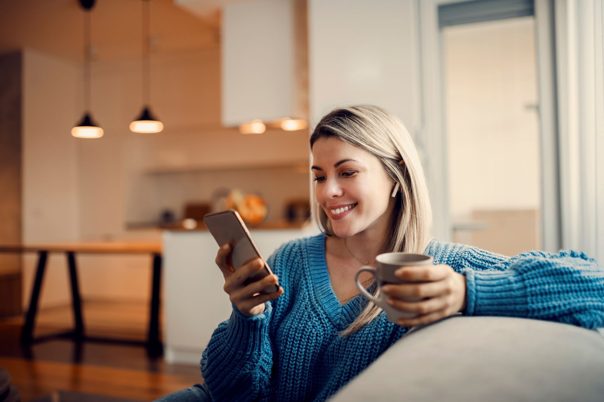 Woman in blue sweater smiles while looking at phone, holding coffee cup, indoors.