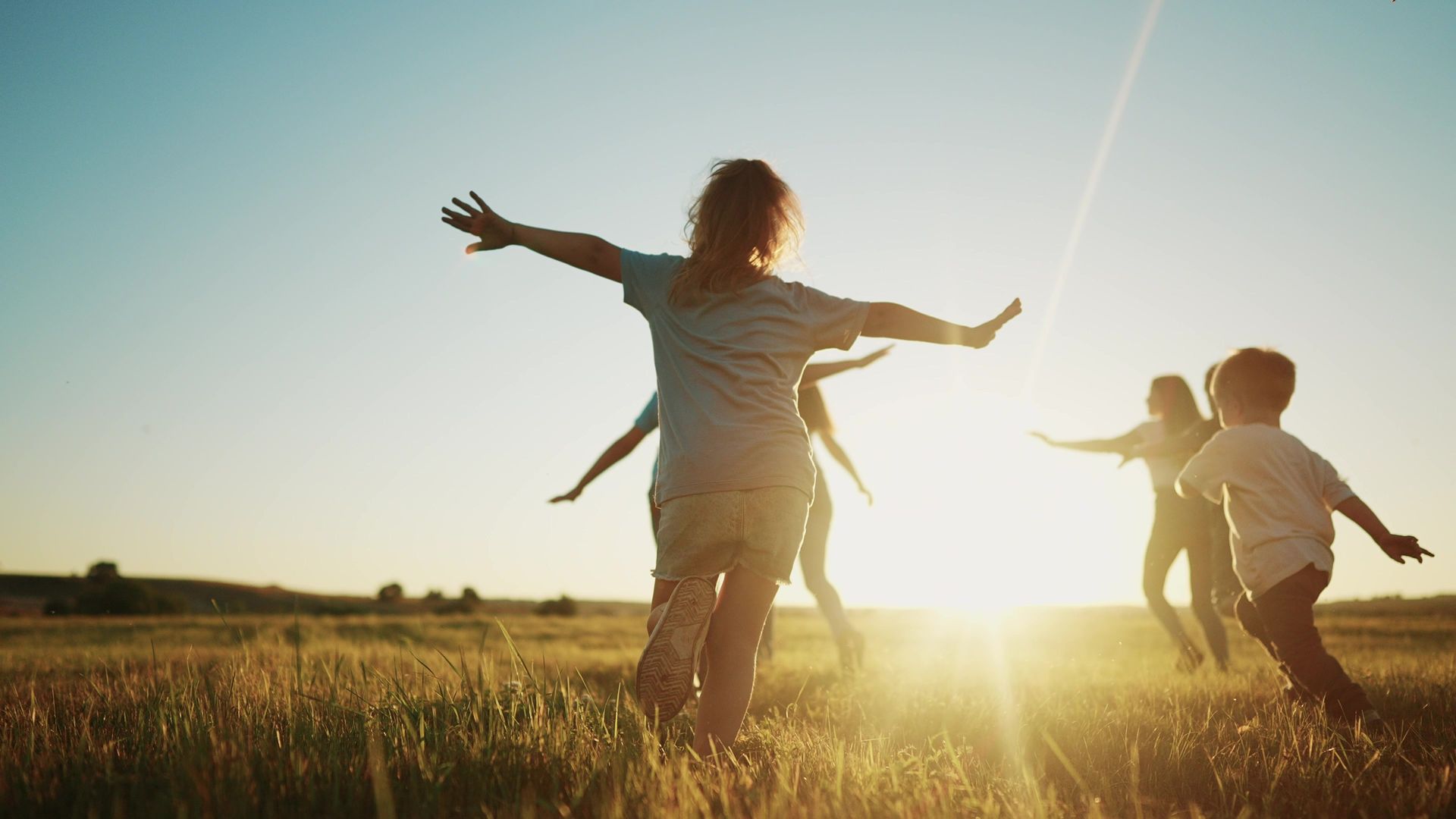 People with arms outstretched, running through tall grass towards setting sun.