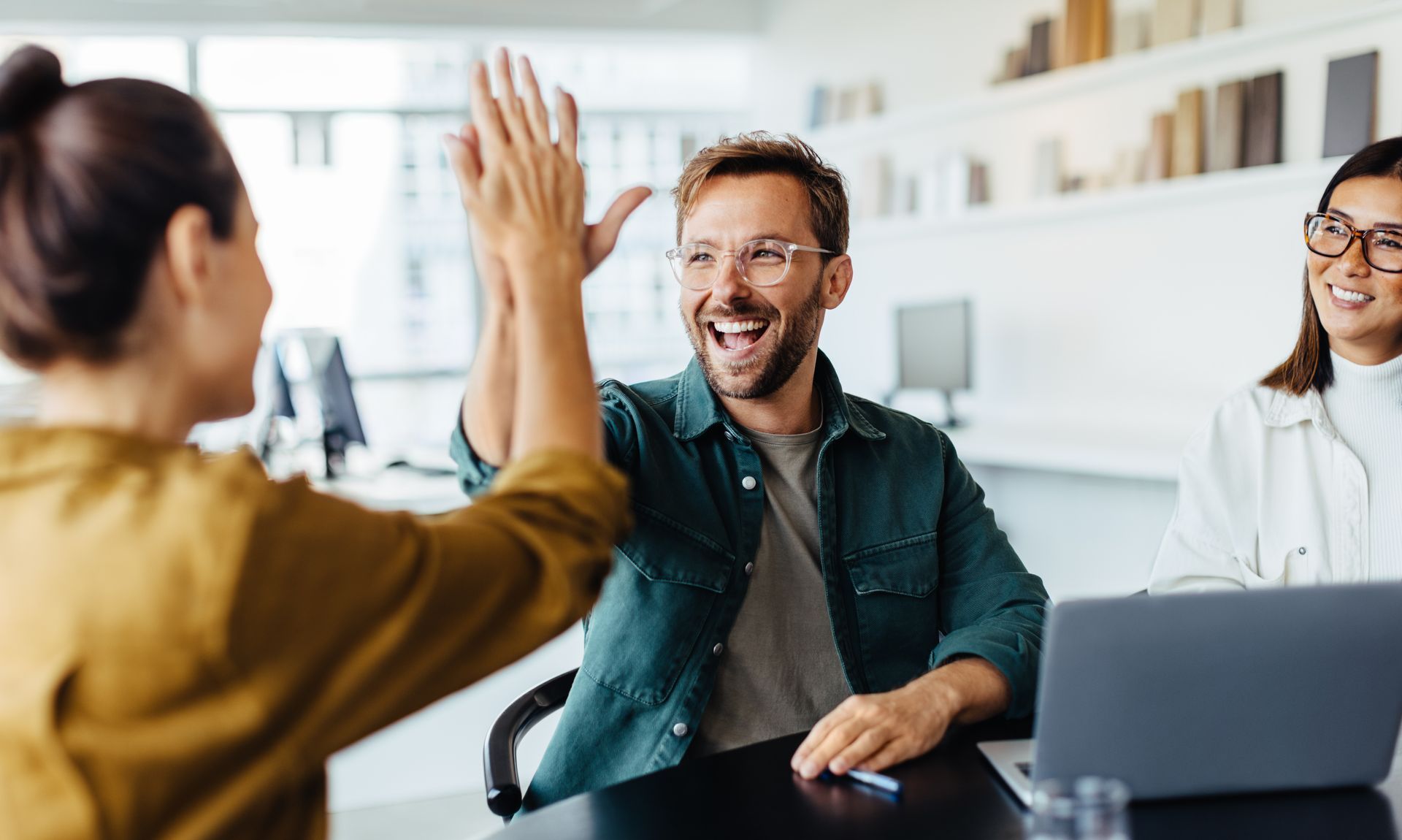People giving a high five in a bright office. Man smiles, excited. Woman wearing glasses smiles as well.