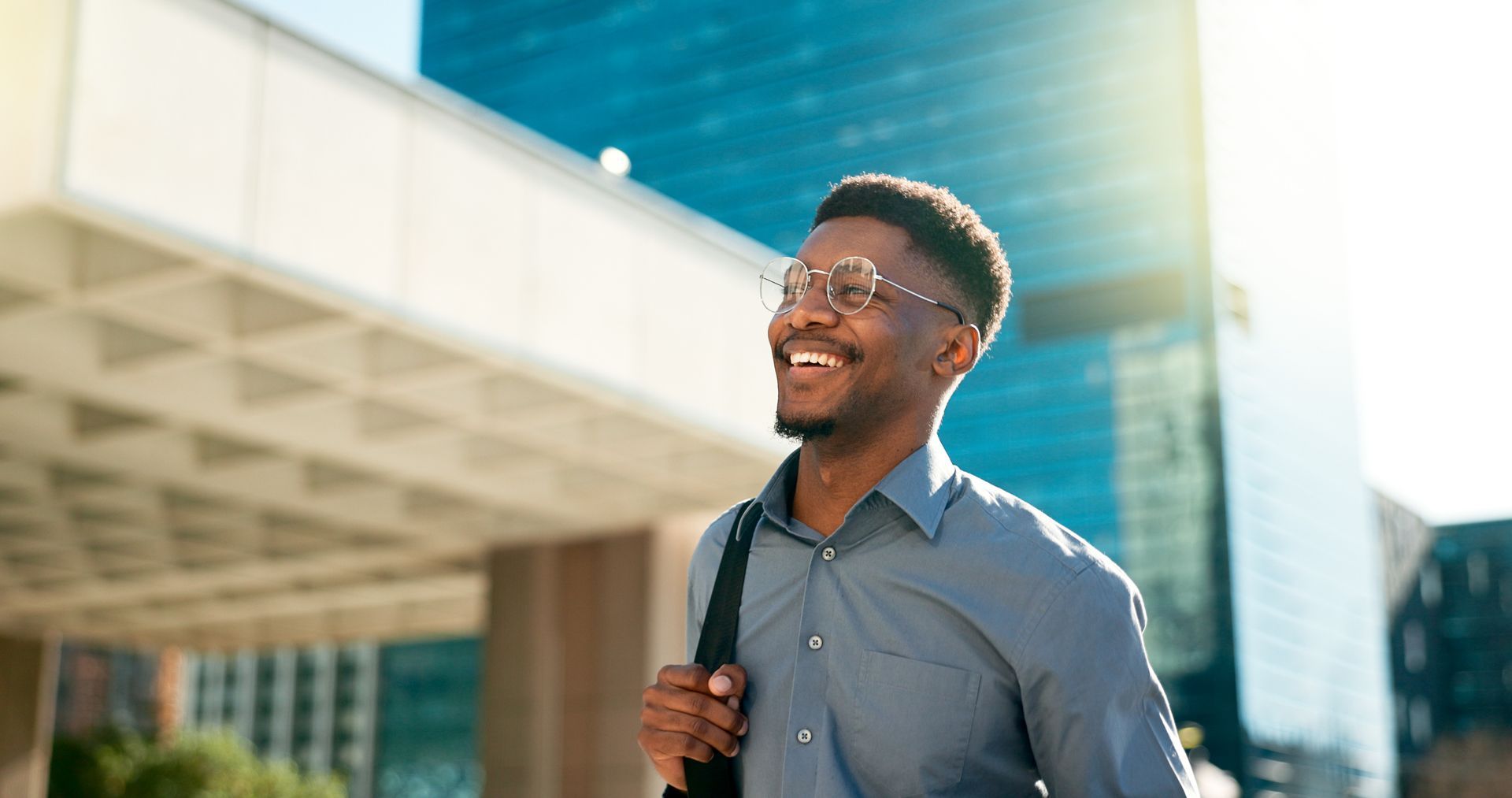 Smiling man walking outside a building, wearing glasses and carrying a bag.