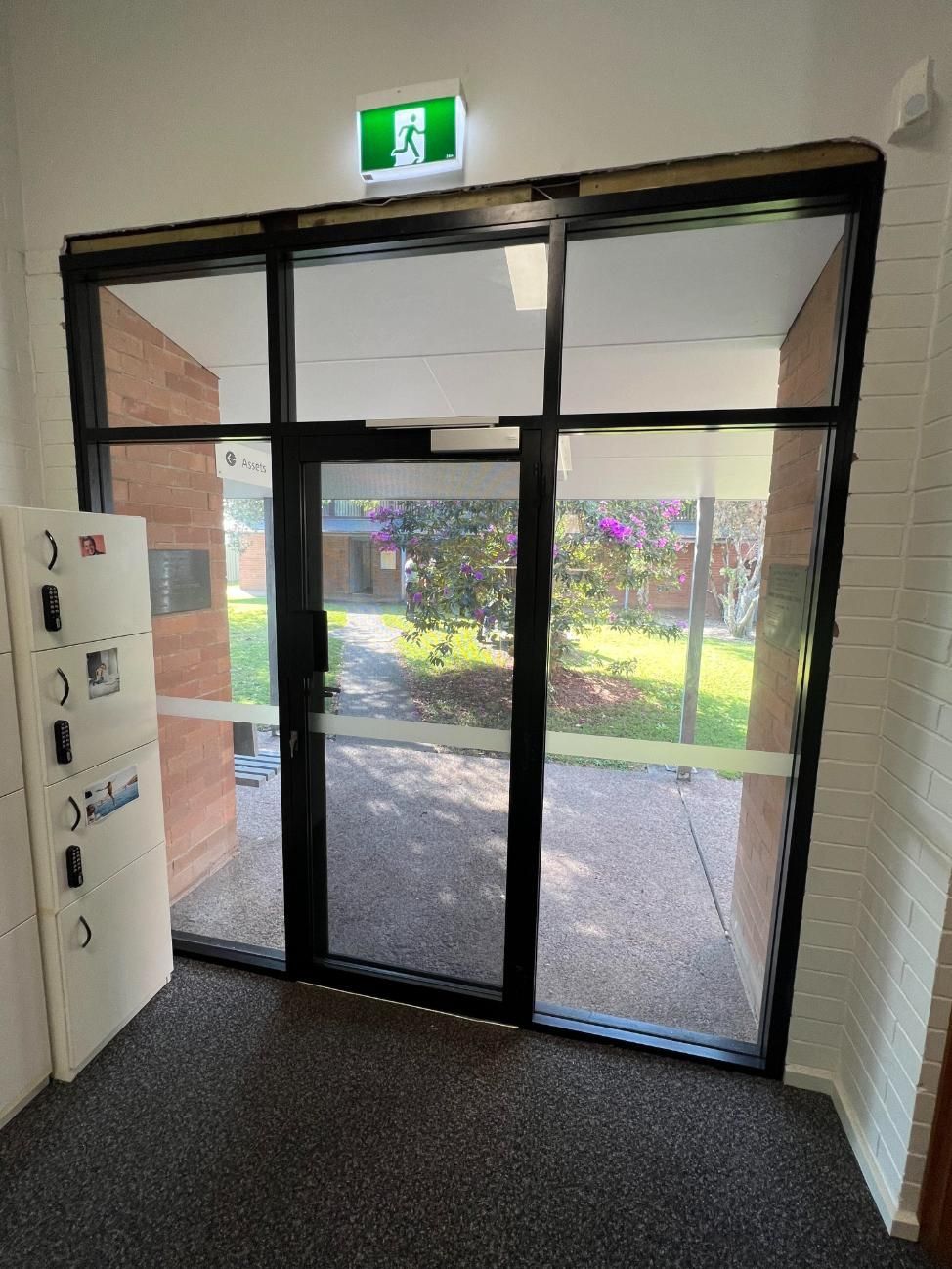 A Hallway With A Glass Door And A Green Exit Sign — Glass 24/7 In Forster, NSW