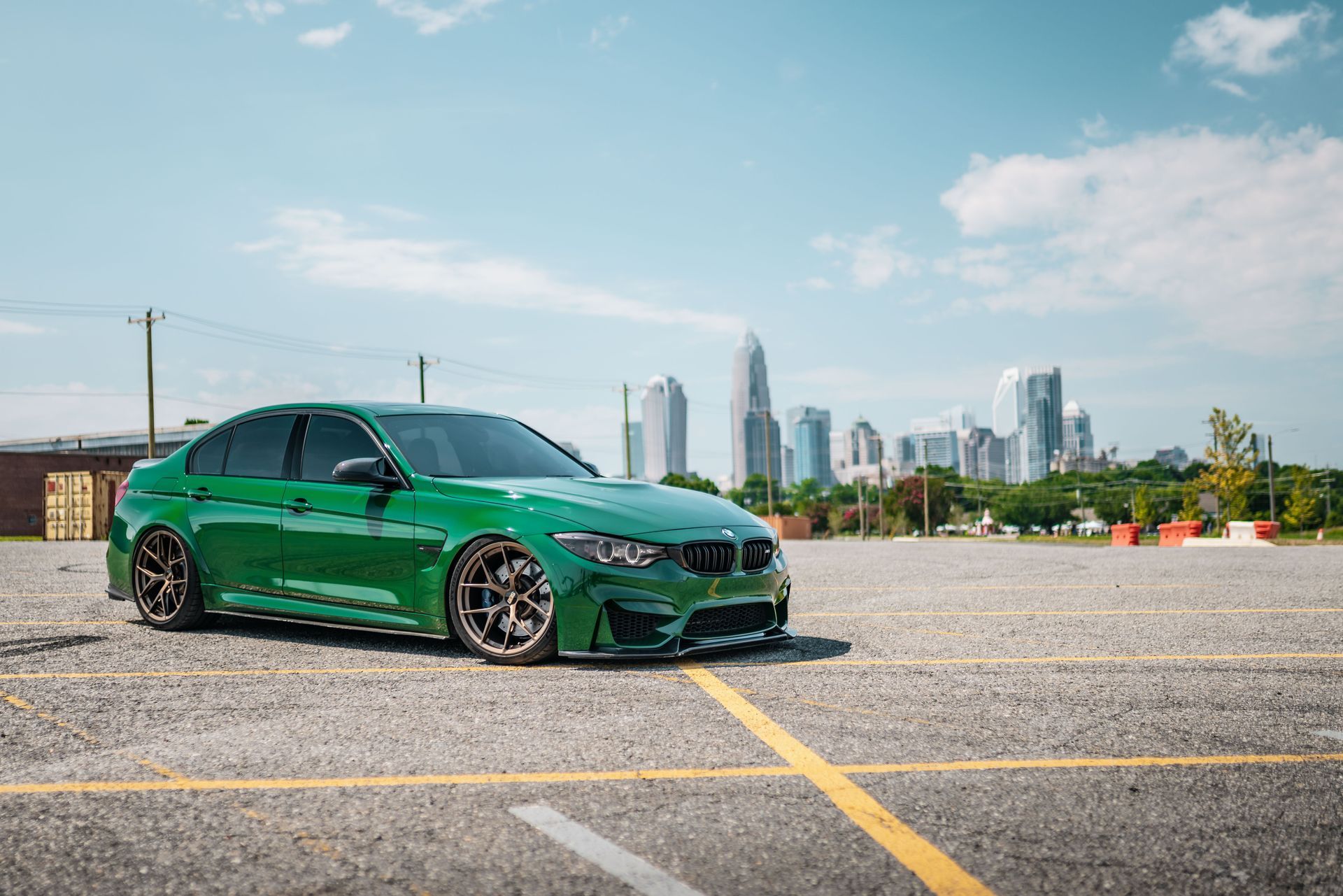 A green bmw m3 is parked in a parking lot with a city skyline in the background.
