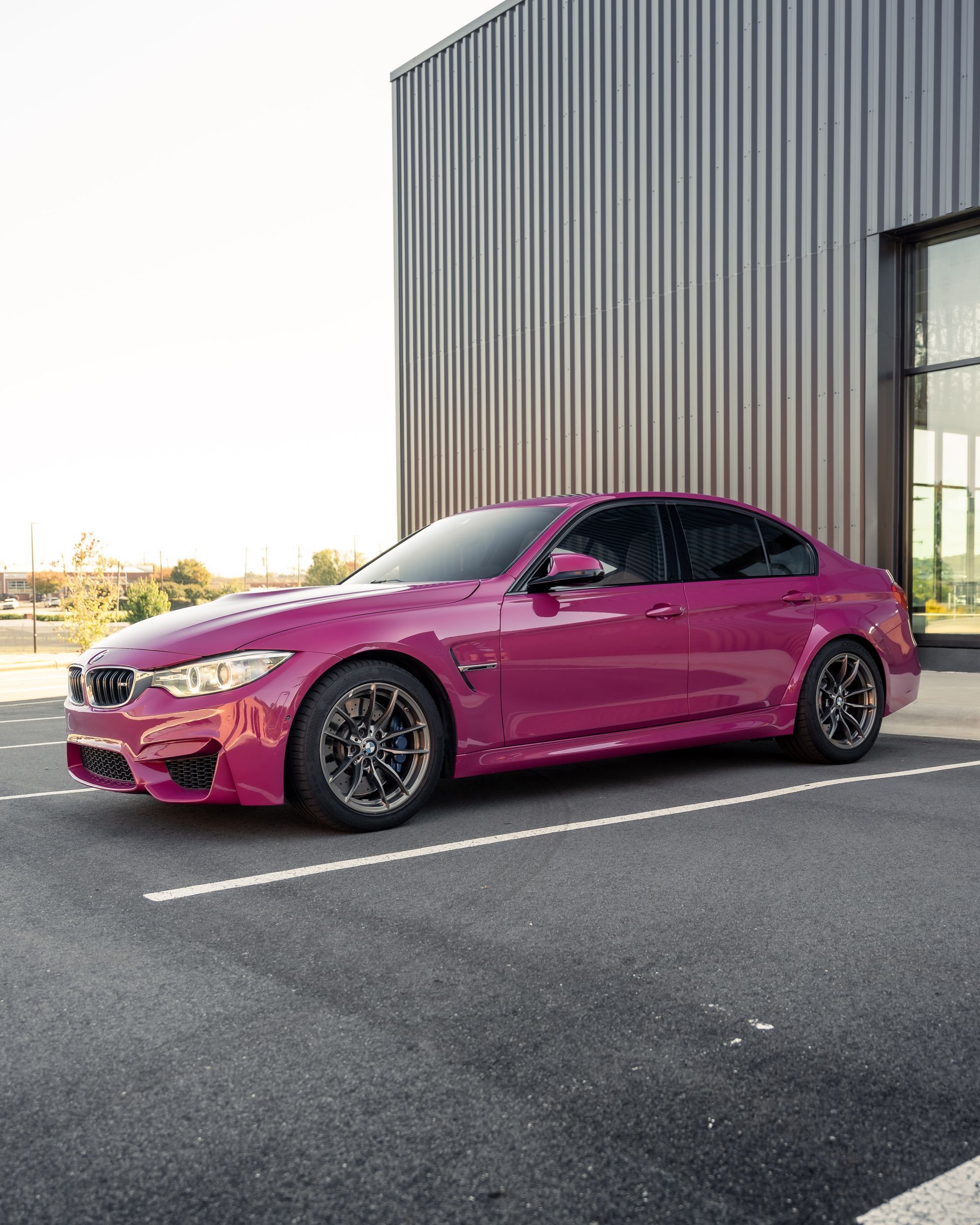 A pink bmw m3 is parked in a parking lot in front of a building.