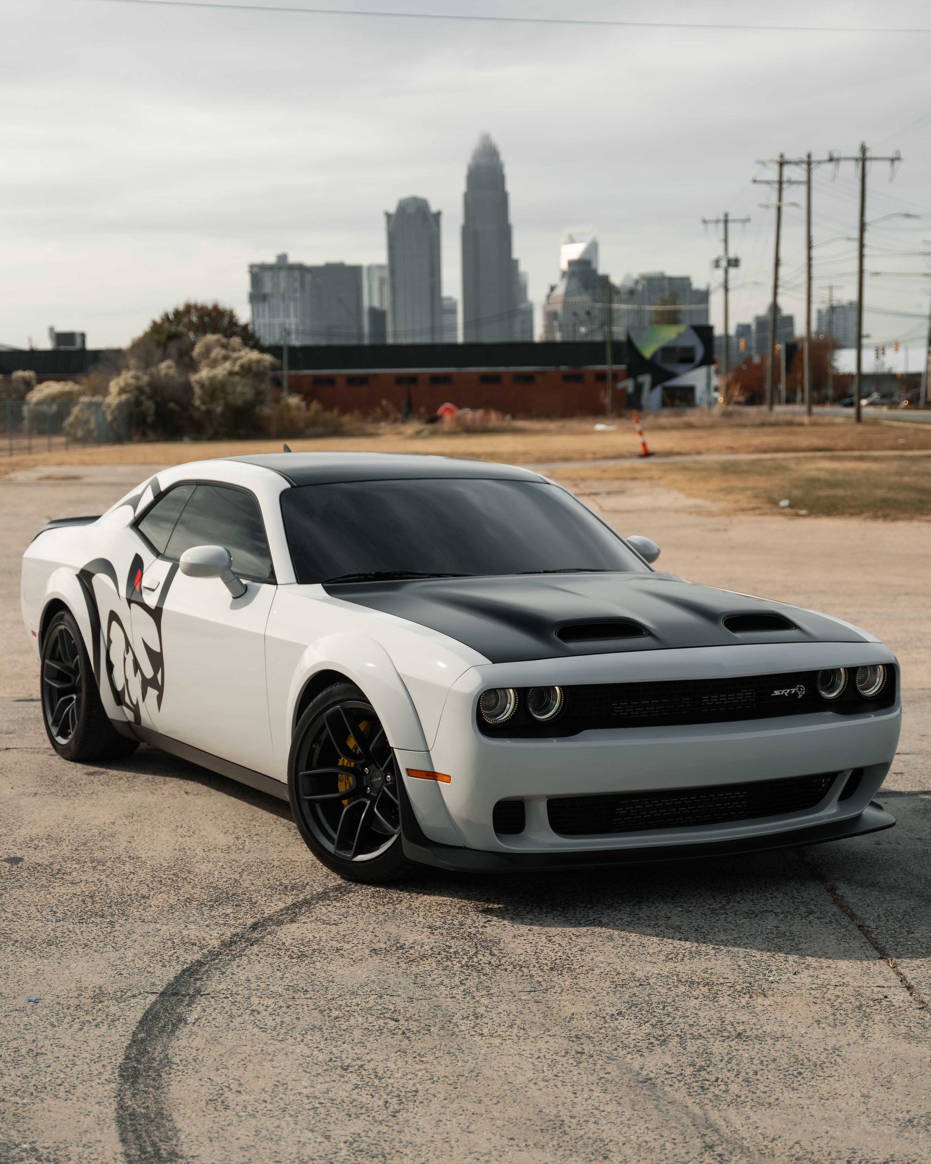 A white dodge challenger is parked in a parking lot in front of a city skyline.