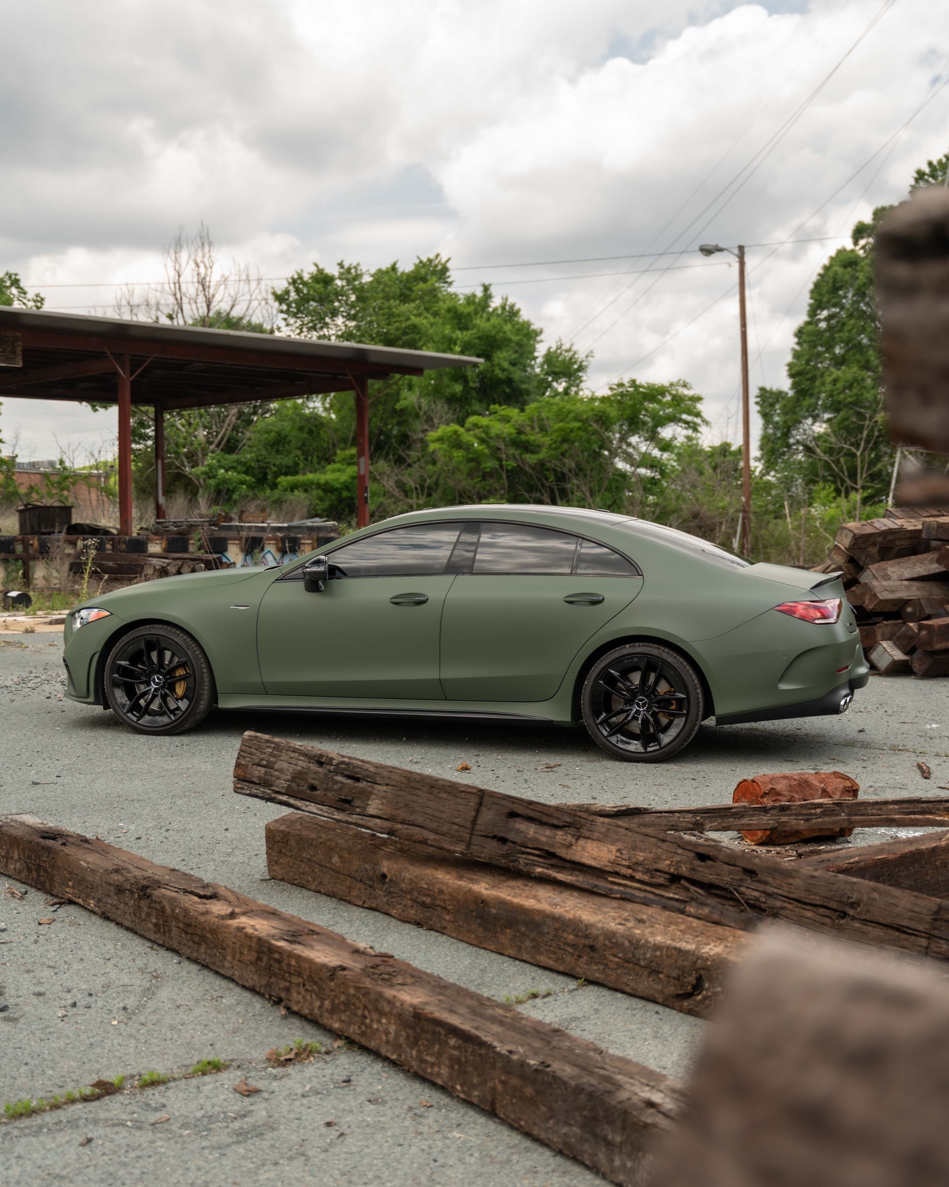 A green car is parked in a parking lot next to a pile of wood.
