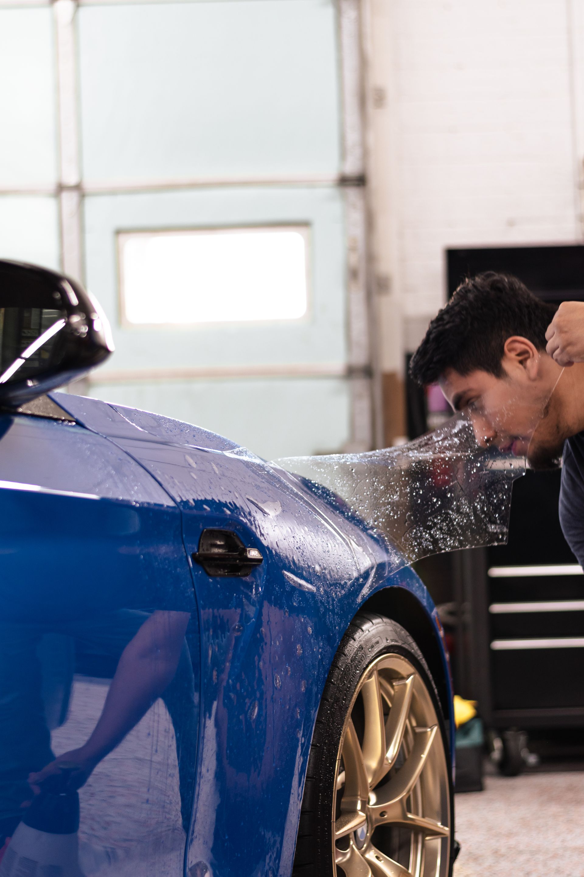 A man is applying a protective film to a blue car in a garage.
