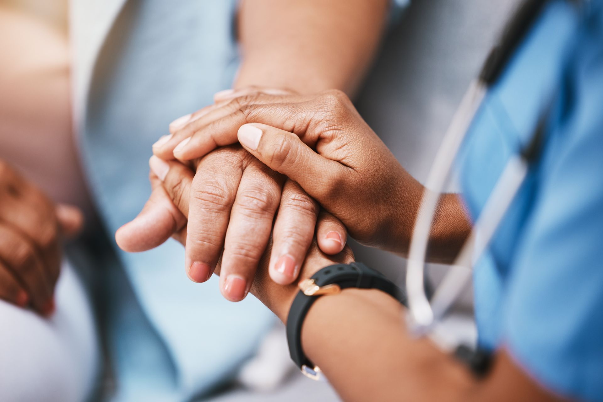 A healthcare worker in blue scrubs holds a patient's hand in a gesture of comfort and support.