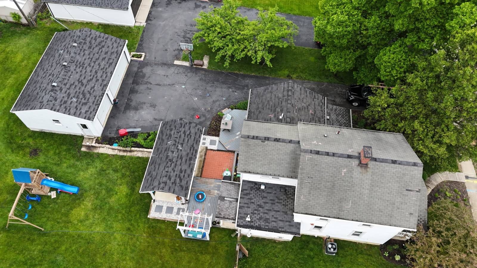 Aerial view of a house with a black roof, driveway, detached garage, and a green lawn.