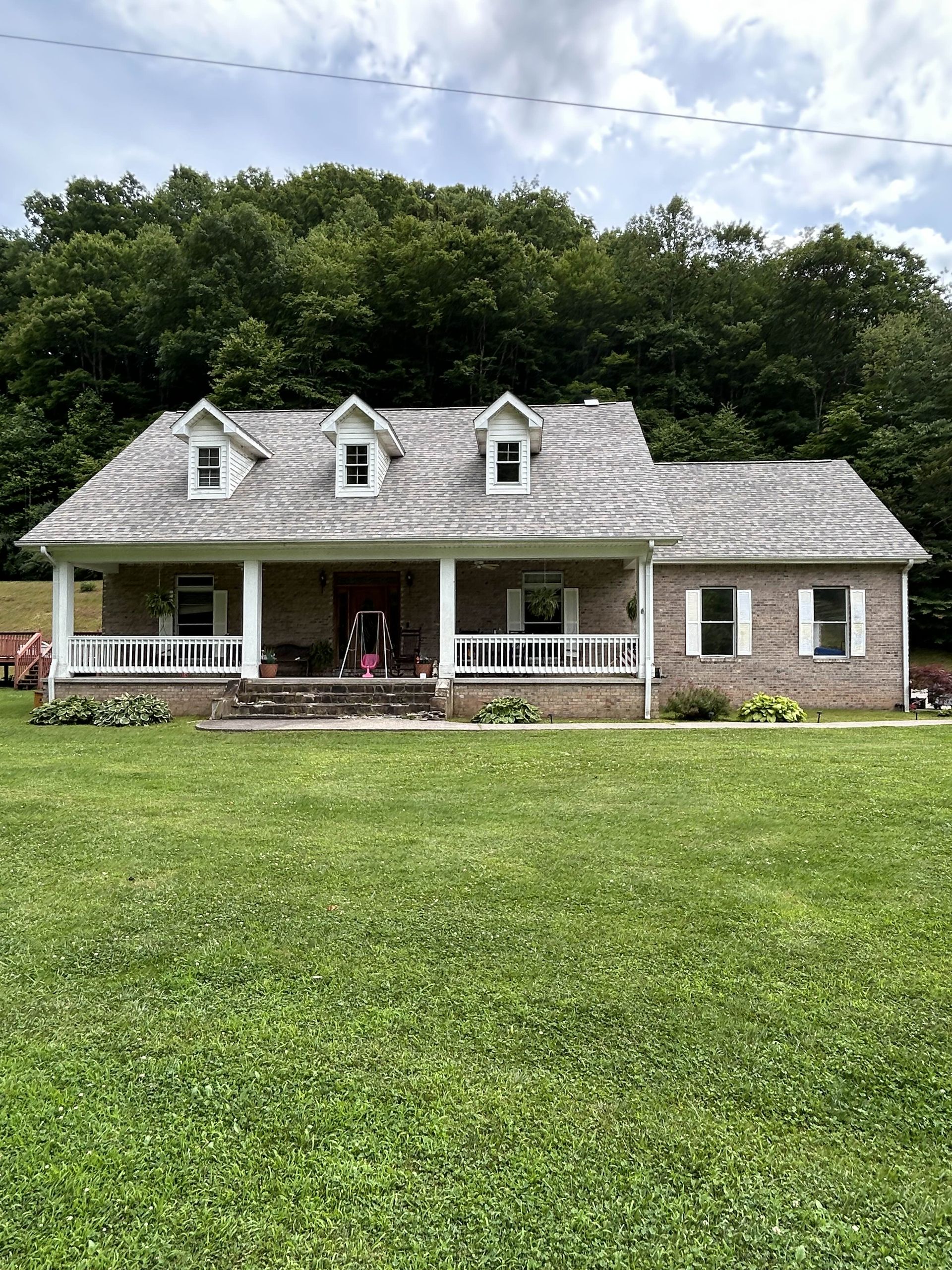 White house with porch, dormers, and stone accents in a grassy yard, backed by trees.