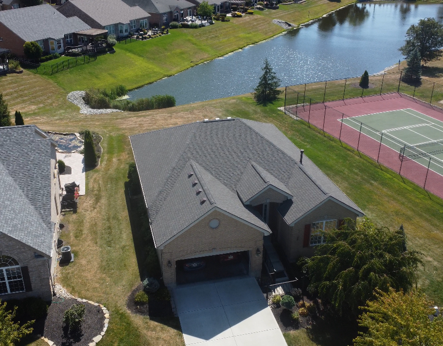 Aerial view of a house with a gray roof and a driveway, near a lake and a tennis court.