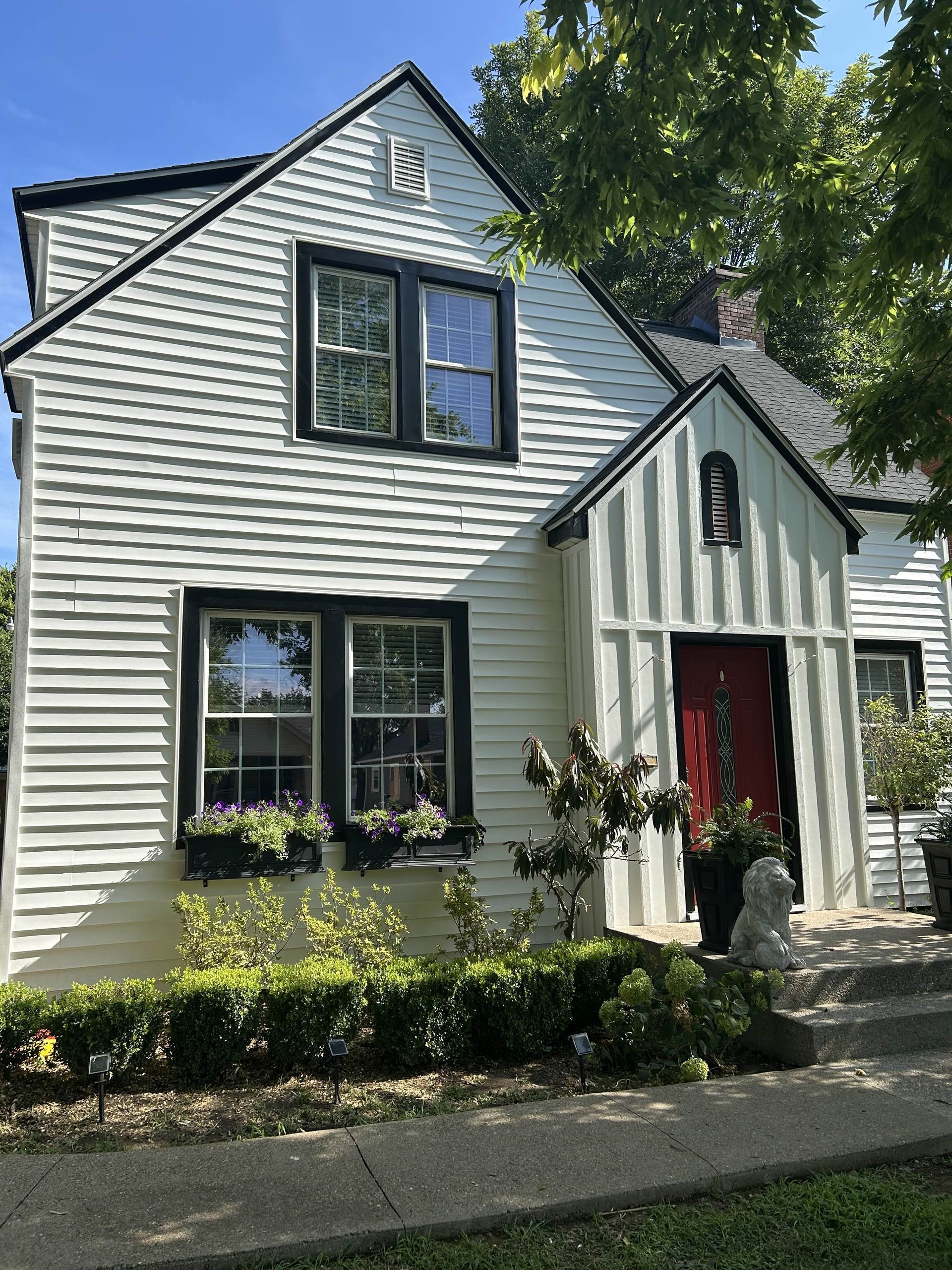 White house with black trim, red door, and flower boxes; trees and bushes in the yard.