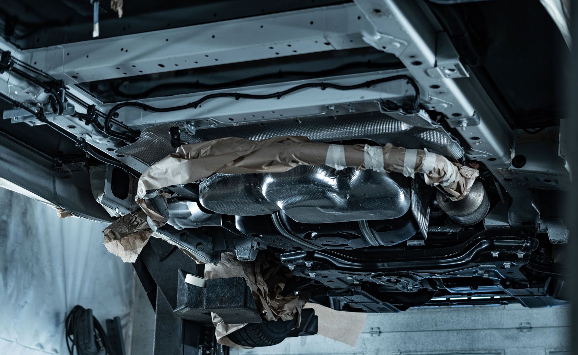Underside of a vehicle frame in a workshop, showing exhaust system, pipes, and protective coverings.