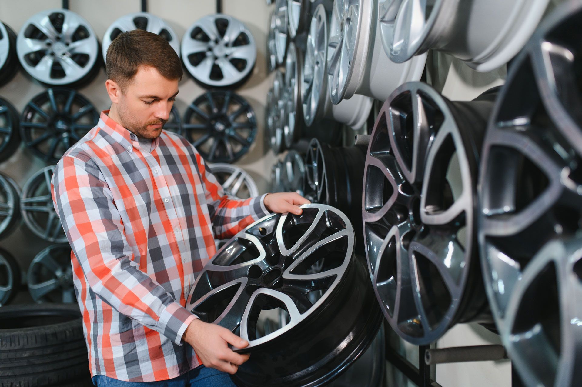 Man in plaid shirt examining a car wheel in a shop filled with wheels.