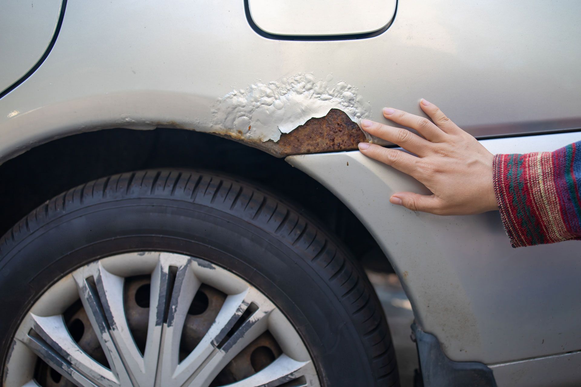 Severely rusted car body, wheel, and person's hand touching the damaged area.