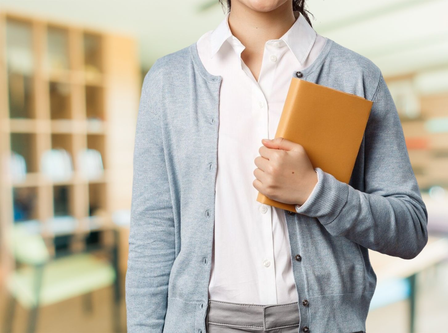 Persona que sostiene un libro, vestida con un cárdigan azul, una camisa blanca y una falda gris, en un entorno de oficina.