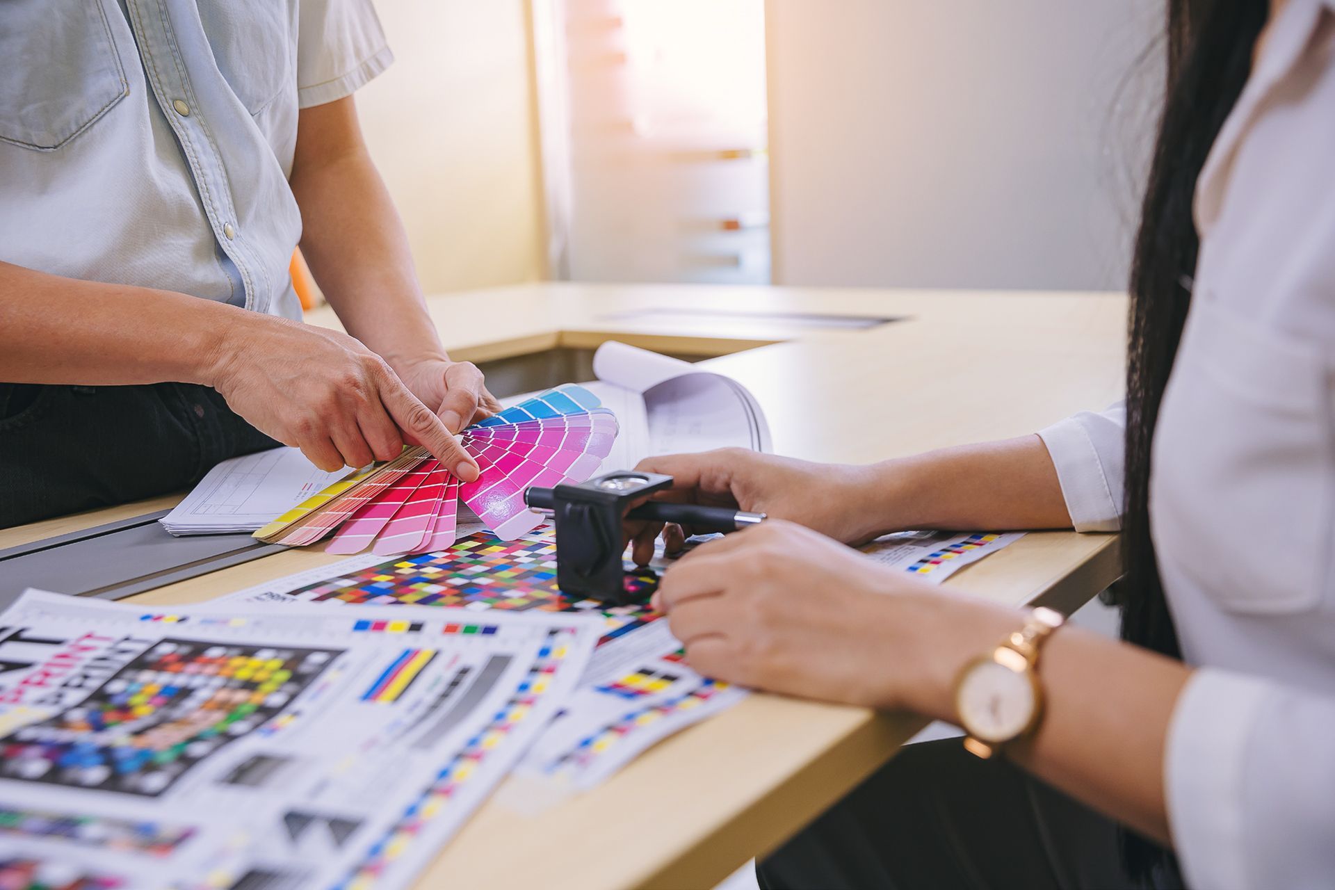 Dos personas revisan muestras de color y de impresión en una mesa en una oficina bien iluminada. Una señala un tono rosa.