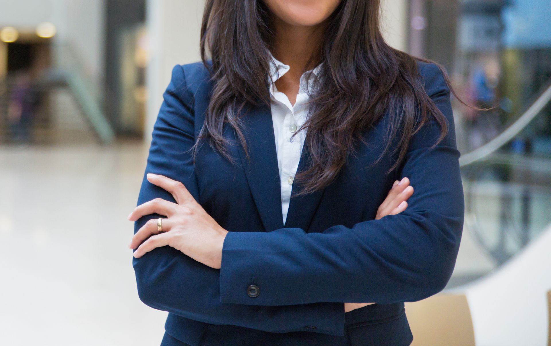 Mujer con blazer azul marino y camisa blanca, con los brazos cruzados, sonriendo levemente, en el interior.