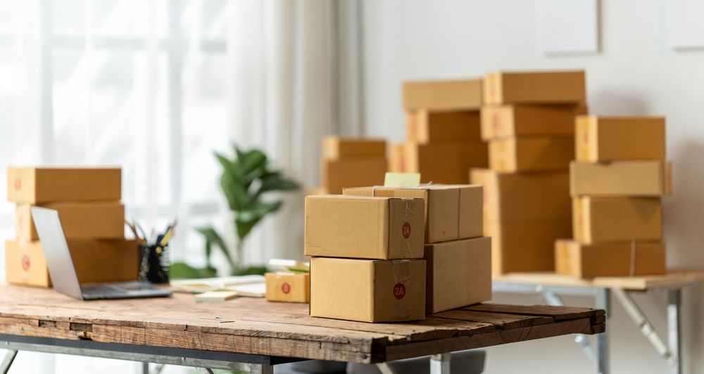 Packed cardboard boxes on a table in a bright room with more stacked boxes in the background